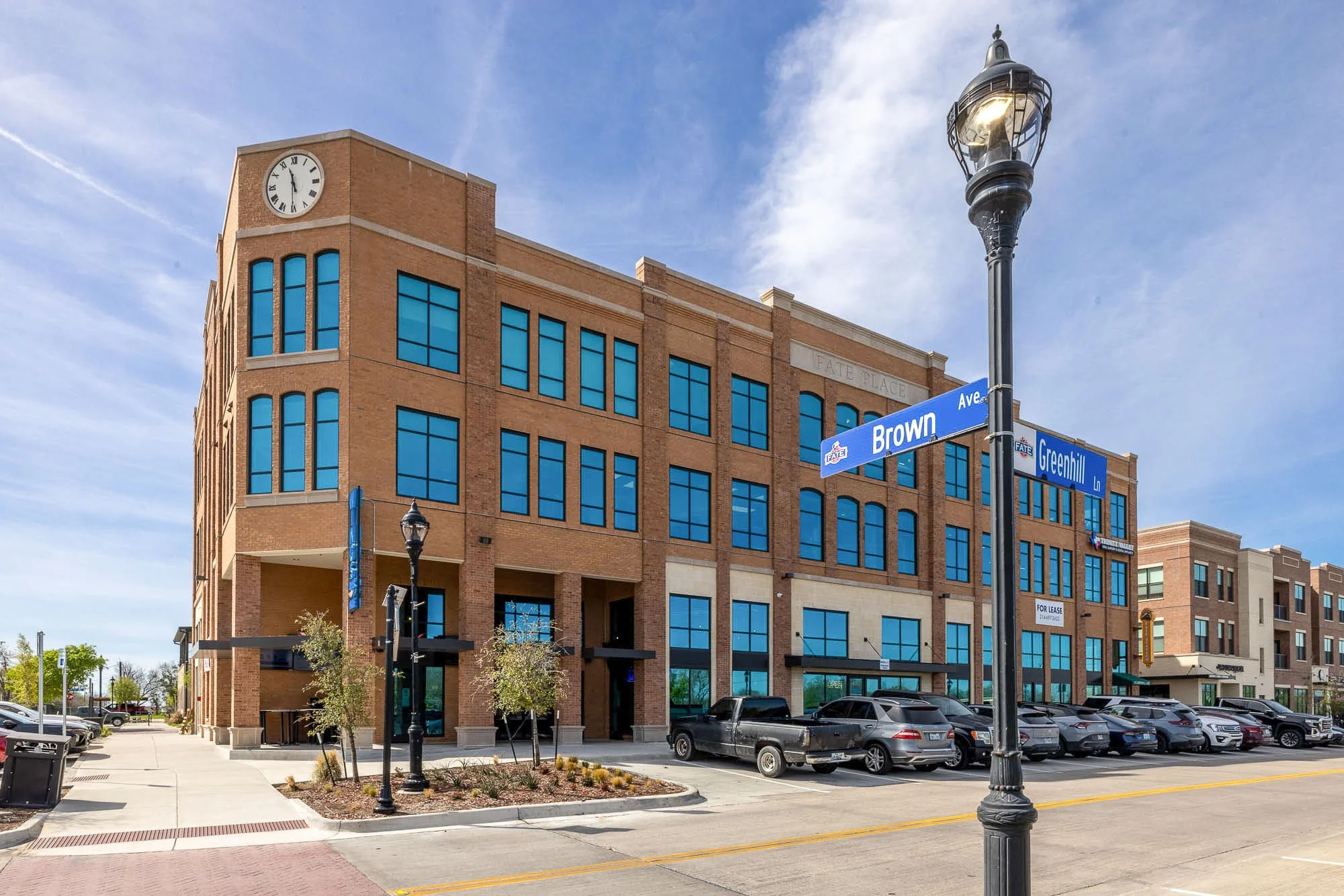 A multi-story brick building with a clock showing 2:02 and large blue-tinted windows, situated along a parking lot with cars. There are street signs indicating Brown Avenue and Greenhill Lane, and a vintage-style street lamp in the foreground. The sky is partly cloudy.