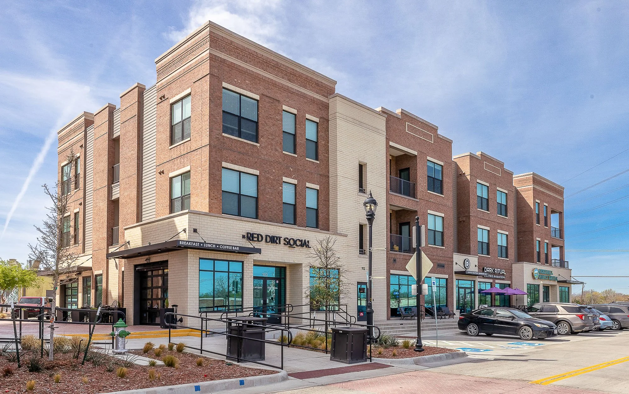 A multi-story commercial building with a sign reading 'Red Dirt Social' and various business logos on the ground floor, a parking lot with cars, and a sidewalk with outdoor seating under purple umbrellas.
