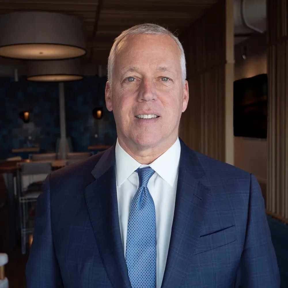 A middle-aged man with short gray hair wearing a dark blue suit, white shirt, and blue tie, standing inside a modern restaurant or lounge area with dim lighting and curtains.