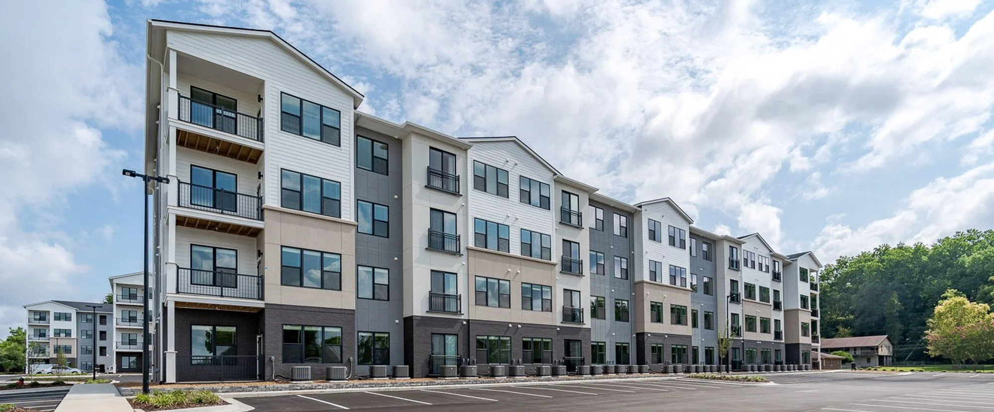Modern multi-story apartment complex with parking lot, trees, and a partly cloudy sky.