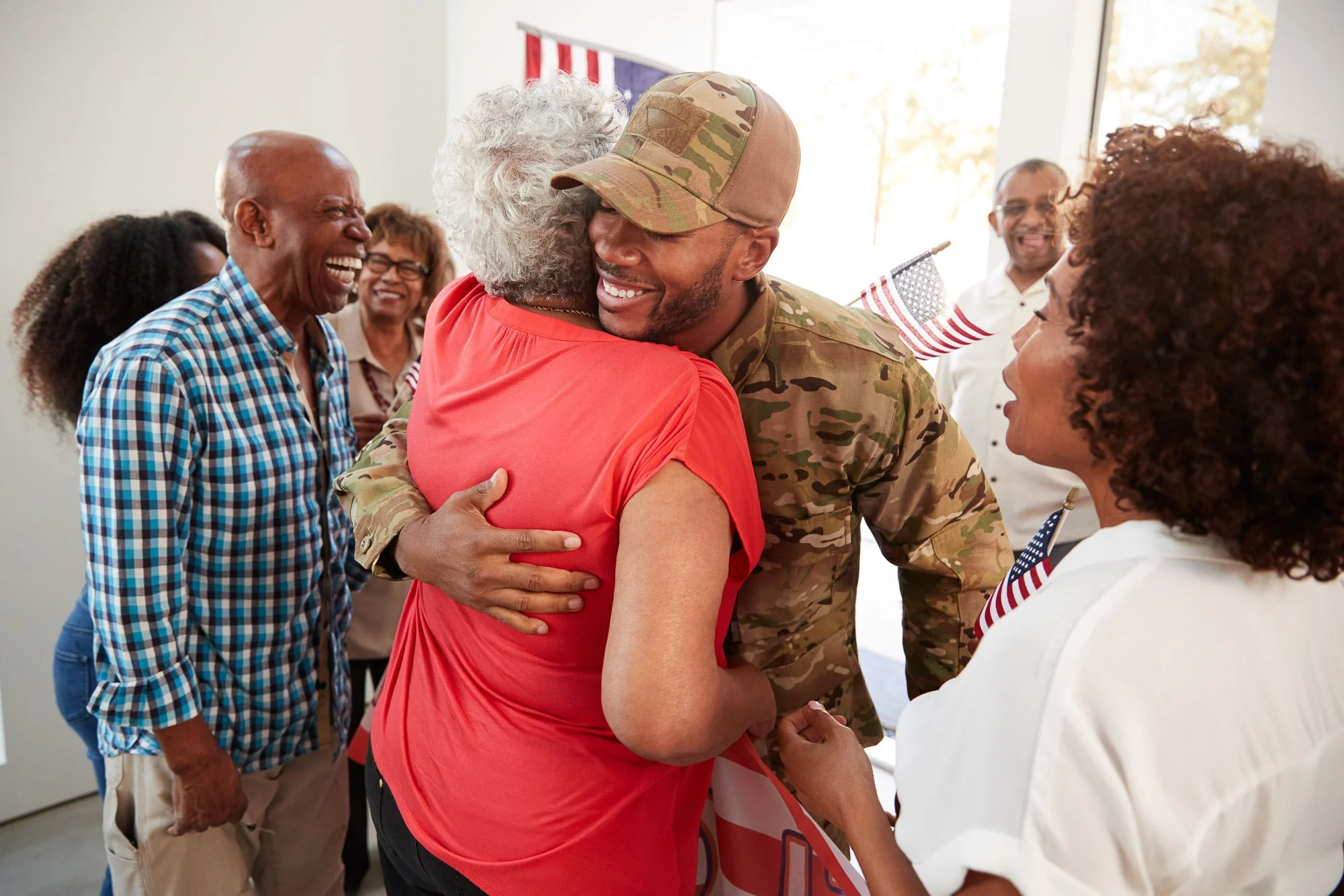 A soldier in camouflage uniform is hugging an elderly woman in a red shirt. They are smiling and appear happy. Several other adults are in the background, smiling and enjoying the moment inside a well-lit room with a window, and an American flag is partially visible on the wall.