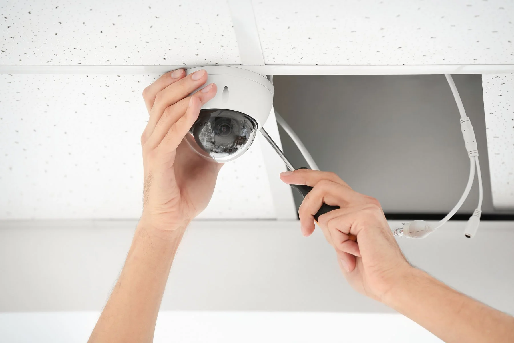 Person installing a security camera on a ceiling with a screwdriver, ceiling tiles and cables visible.