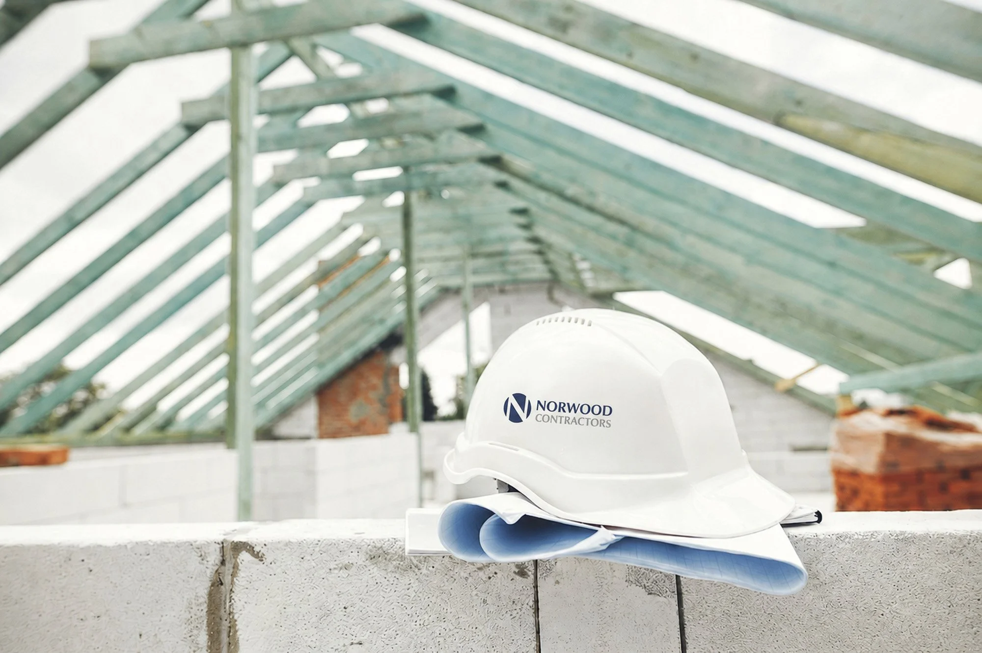 Construction site with green wooden roof structure, white hard hat with "Norwood Contractors" logo, blueprints, and papers on a concrete ledge.