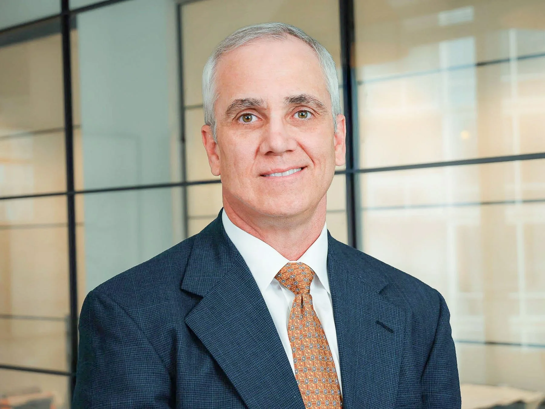 A middle-aged man with gray hair wearing a dark blue suit, white shirt, and orange patterned tie standing in front of a modern glass wall in an office setting.