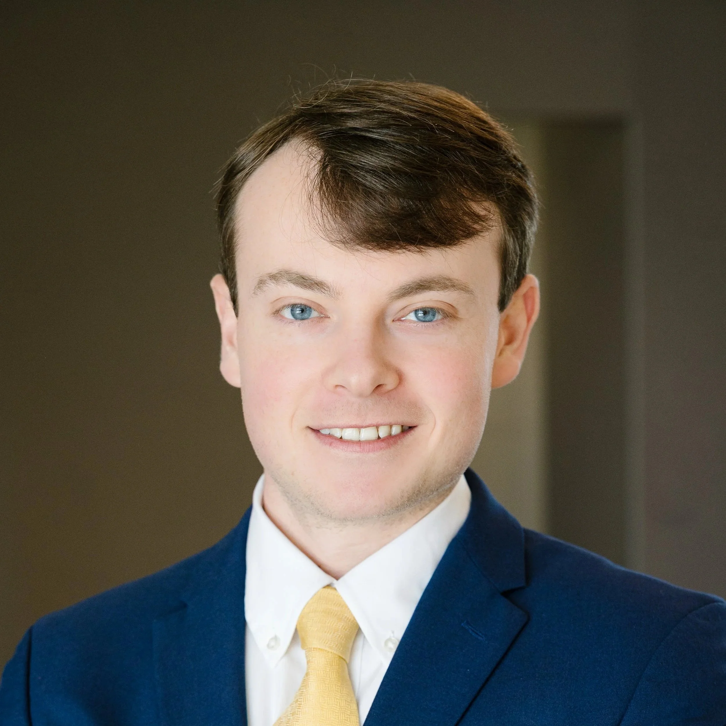 Close-up of a young man with short brown hair, blue eyes, wearing a dark blue suit, white shirt, and yellow tie.