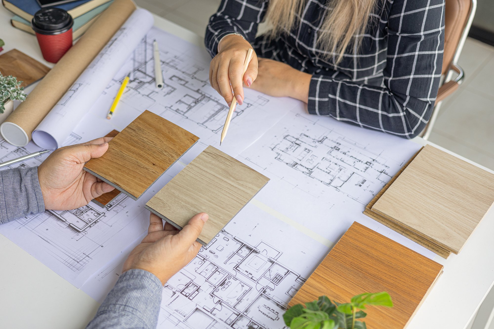 Two people reviewing wooden flooring samples over architectural blueprints, with pens, rolled-up blueprints, and a coffee cup on the table.
