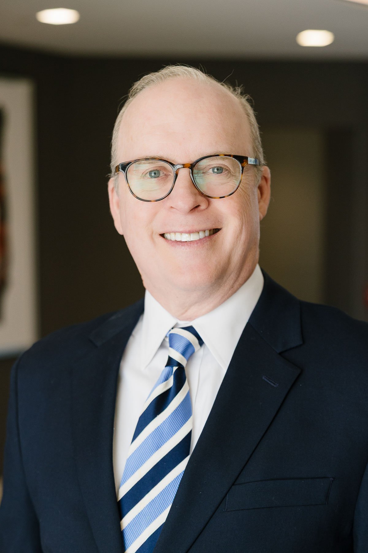 A middle-aged man with light skin, glasses, and a short haircut, smiling in a professional setting. He is dressed in a dark suit, white shirt, and striped tie.