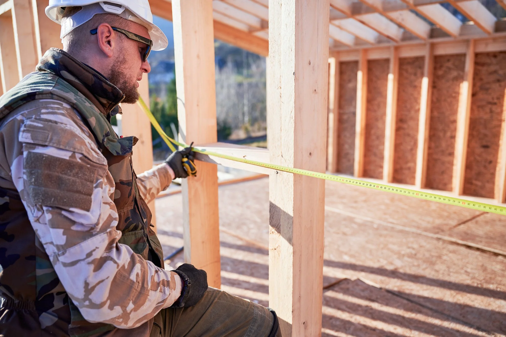 A construction worker wearing a hard hat, sunglasses, and camouflage jacket measures a wooden wall frame with a tape measure at a construction site with exposed wooden beams and a brick wall in the background.