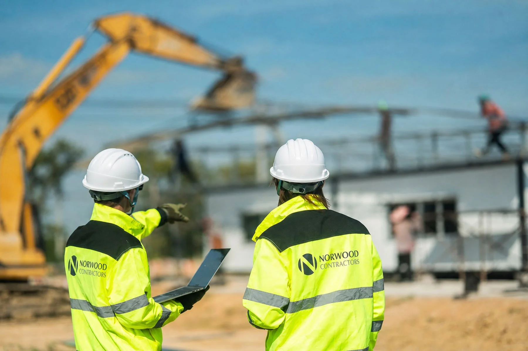 Two construction workers in high-visibility jackets and white hard hats at a construction site, reviewing plans on a tablet, with a crane and construction activity in the background.