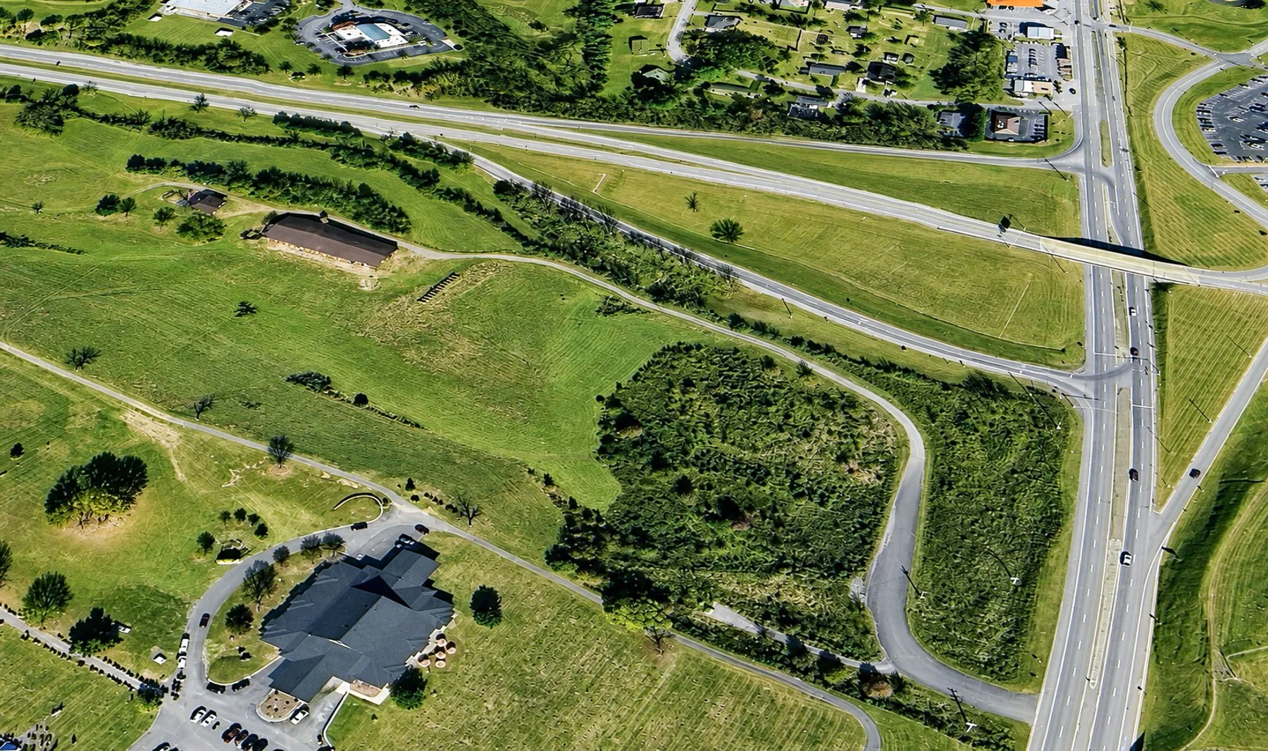 Aerial view of a green park with winding pathways, trees, a building, and nearby roads with cars.