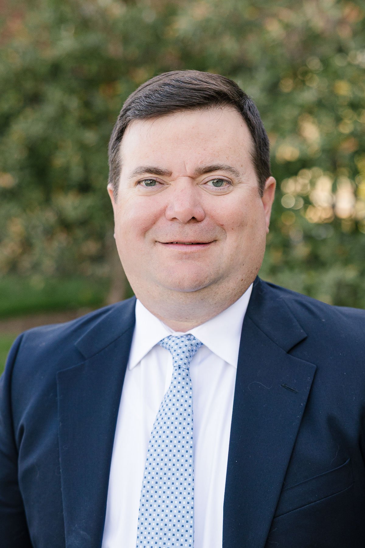 Headshot of a man wearing a navy blue suit, white shirt, and light blue tie with a dotted pattern, outdoors with blurred trees in the background.