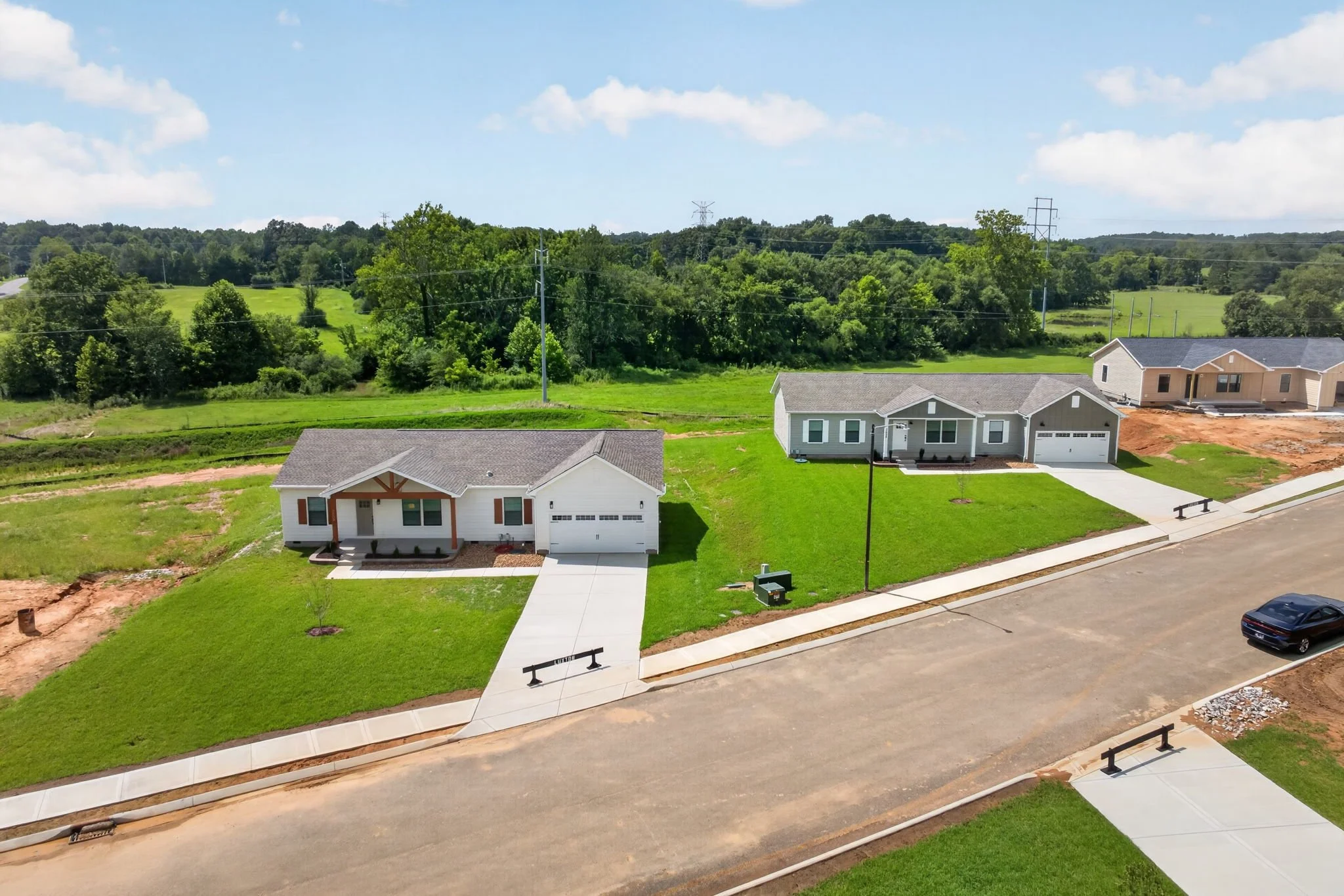 A suburban neighborhood with newly built houses, green lawns, and a paved road and sidewalk, surrounded by trees and a partly cloudy sky.
