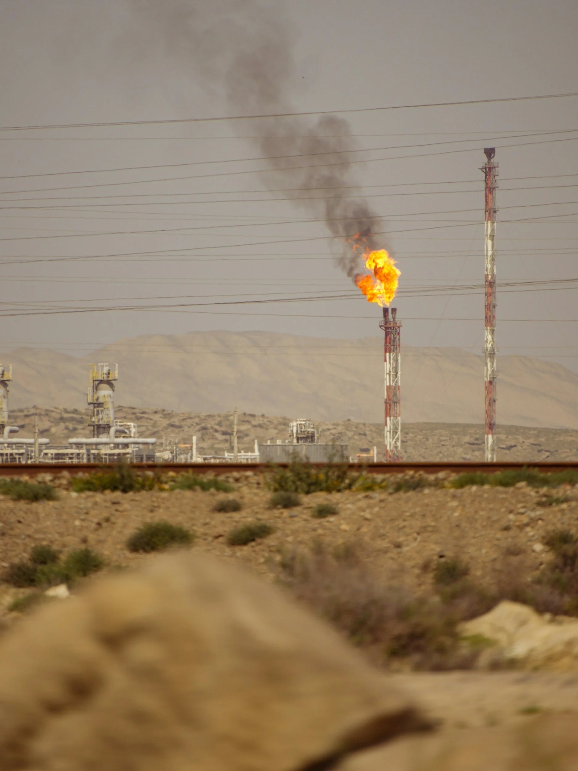 A desert landscape with oil or gas flare stacks emitting flames and black smoke, with power lines crossing the sky.