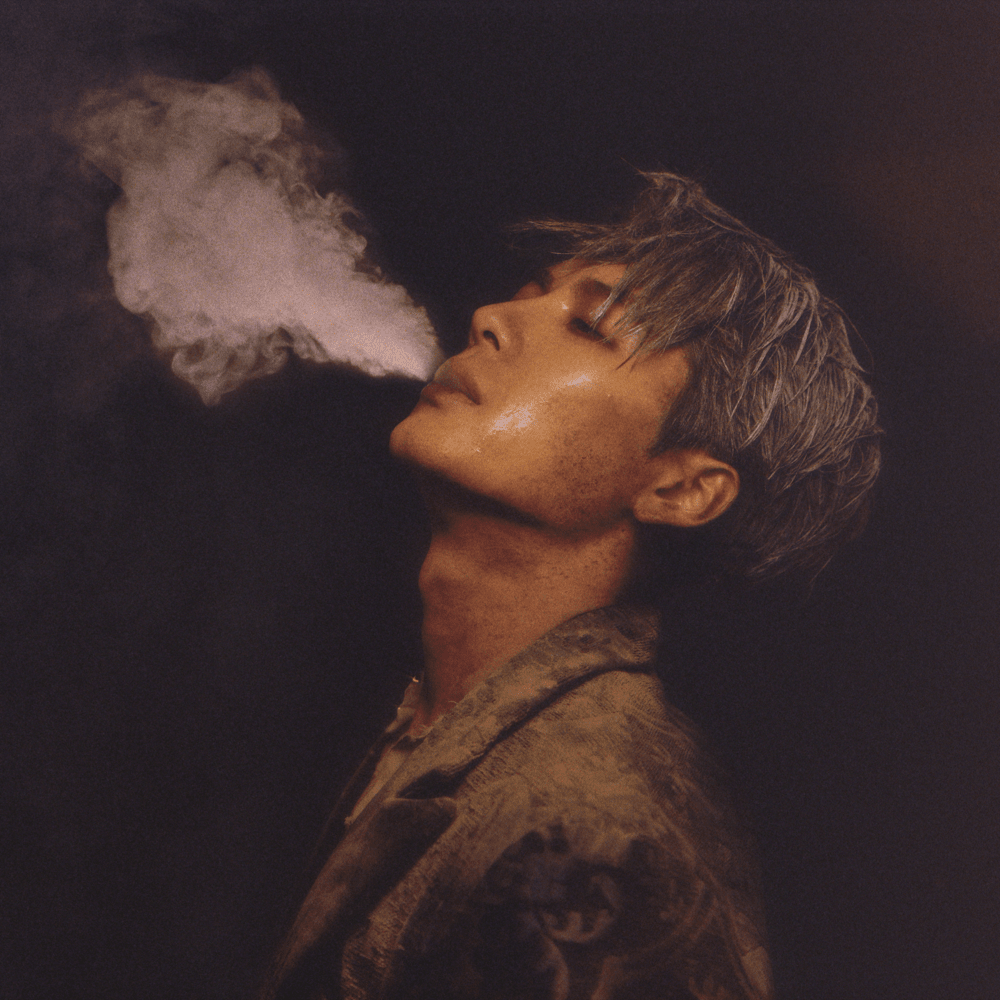 Jackson Wang with grayish hair and freckles exhales smoke from his nose, with his head tilted back against a dark background.