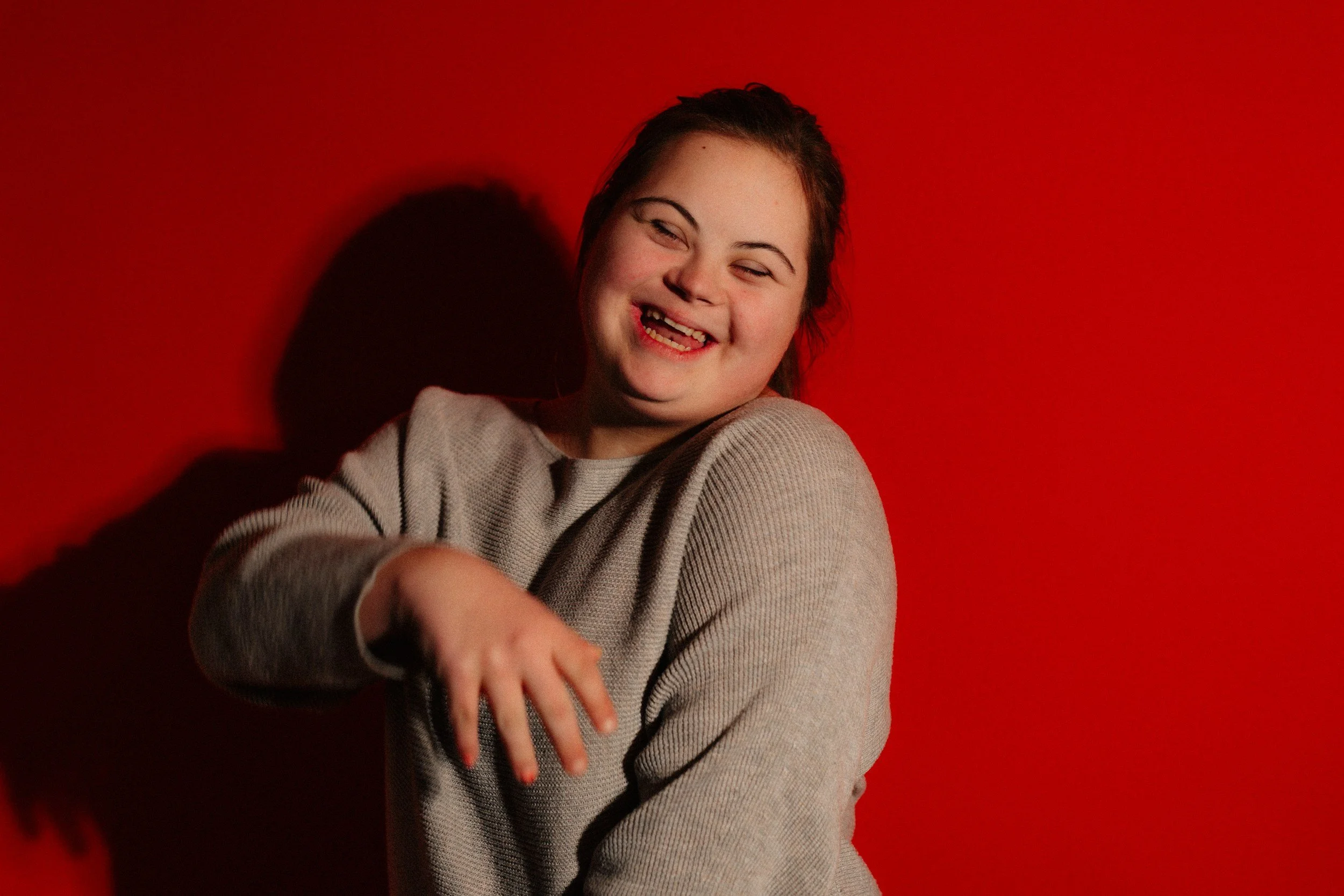 A young developmentally disabled girl smiling and leaning on a red wall with her arm extended toward the camera.