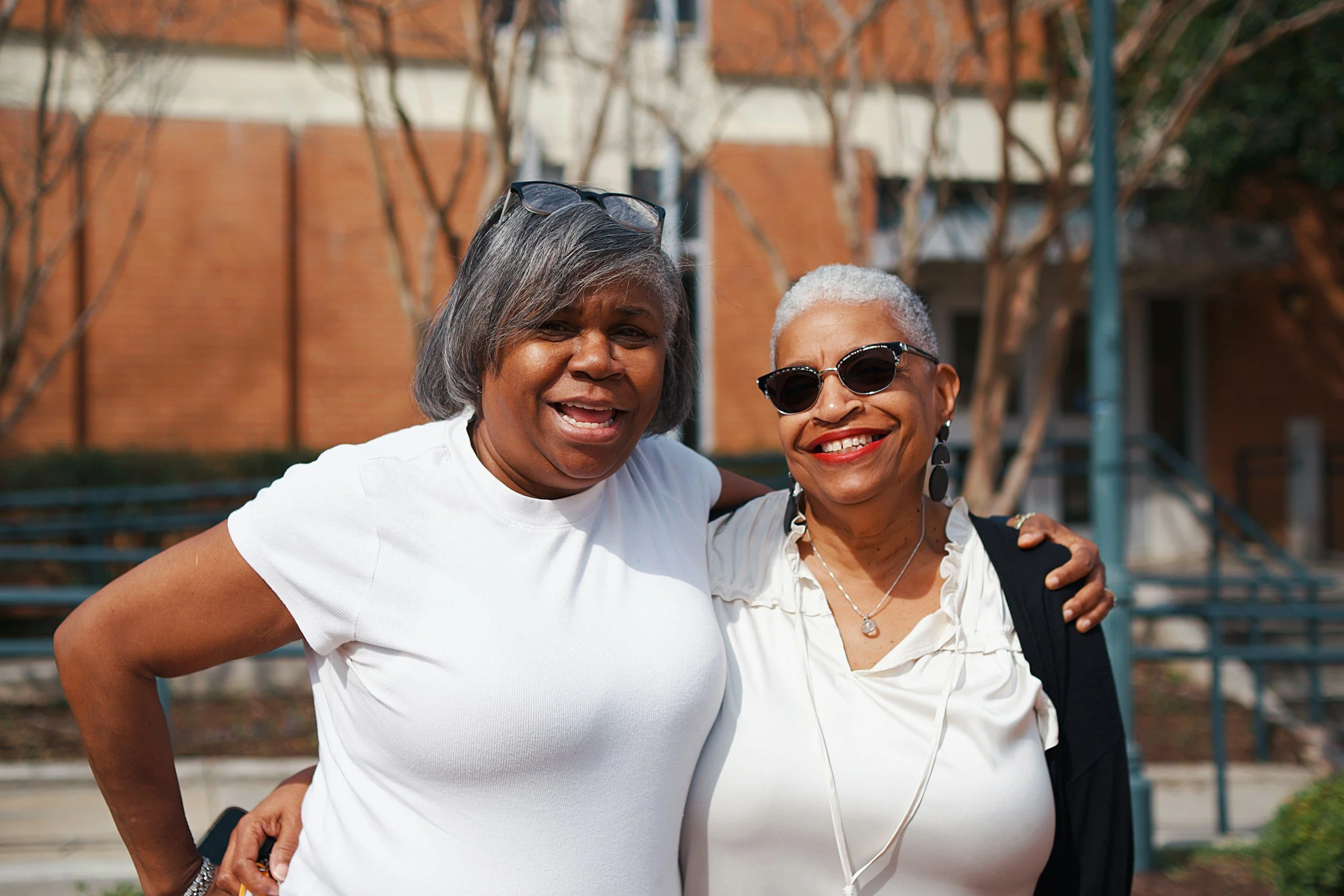 Two women (a mother and daughter) smiling and standing close together outdoors, with one wearing sunglasses and the other with glasses on her head, in front of a background with trees and building structures.