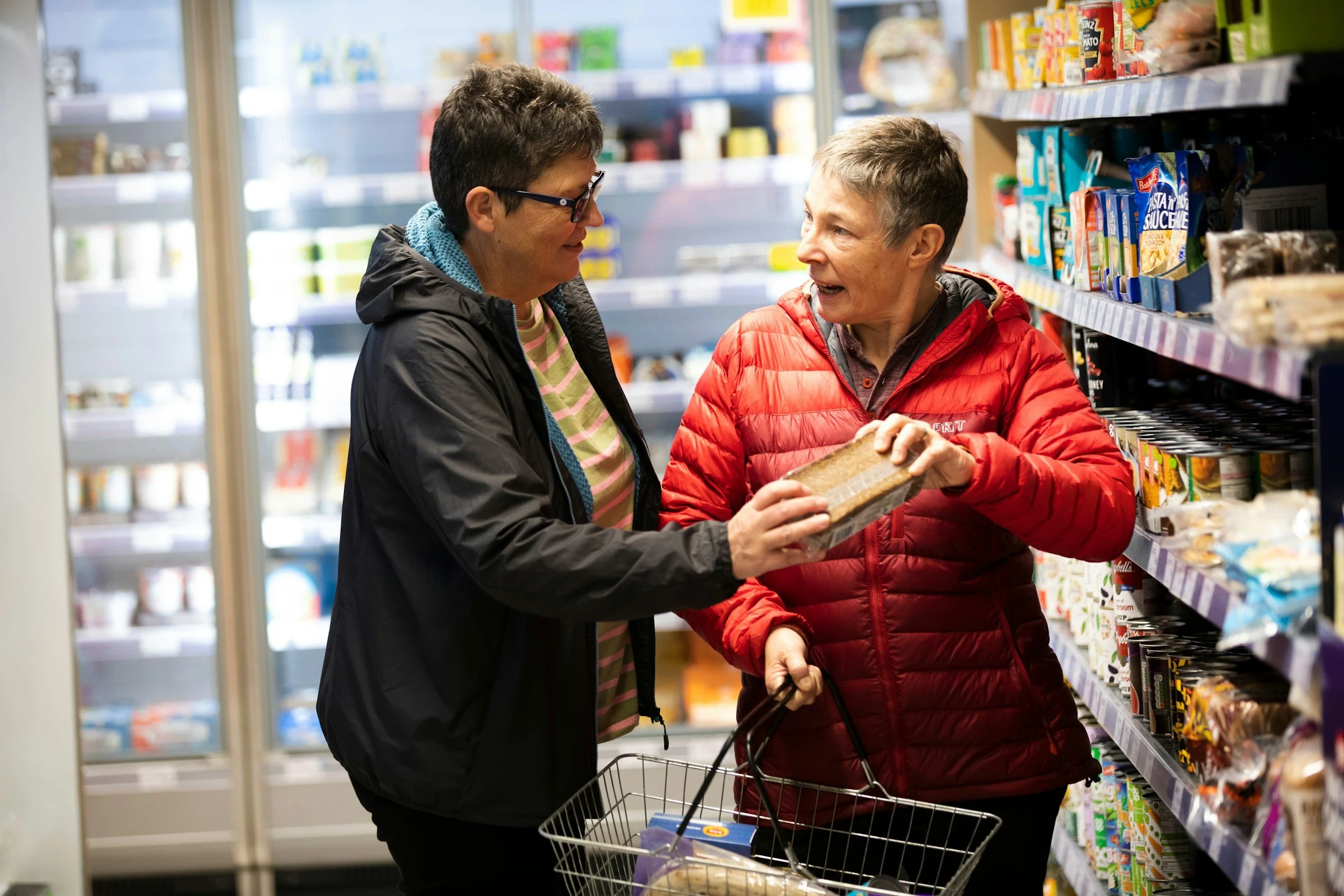 Two women shopping in a grocery store aisle, one woman is supporting the other, handing the other a package of food, both smiling and talking. Teaching daily tasks while providing shopping support.