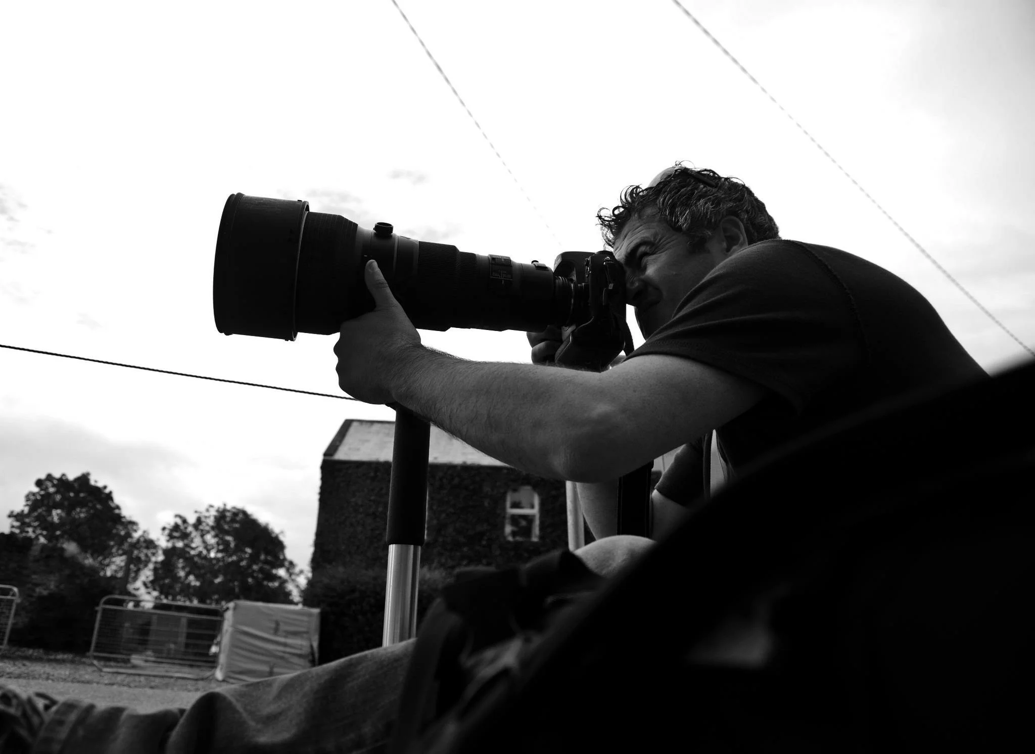 A man looks through a large camera lens, outdoors during daytime, with trees and some structures in the background.