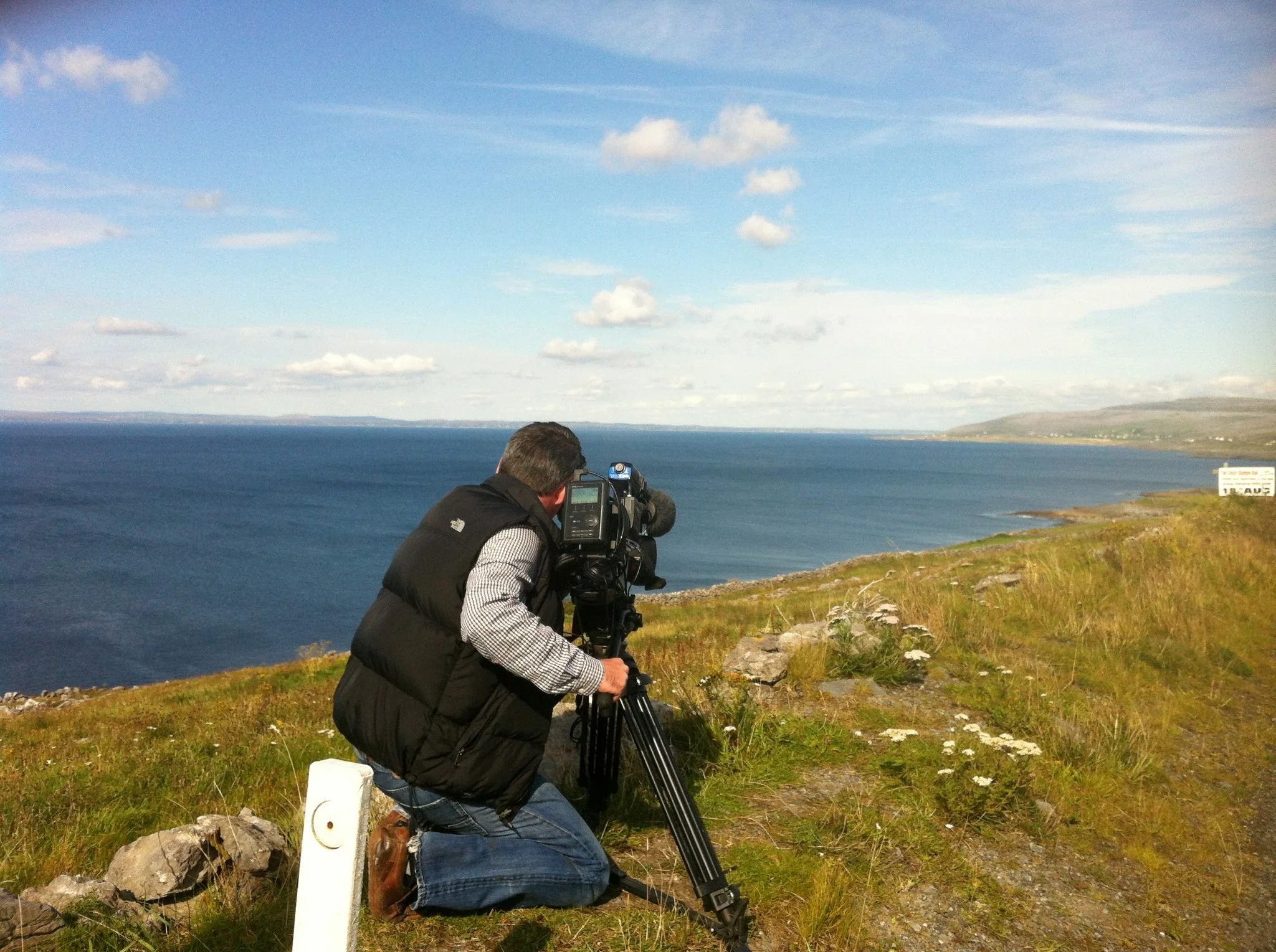 A man kneeling on grass near the coast, operating a professional video camera on a tripod, with ocean and cloudy sky in the background.