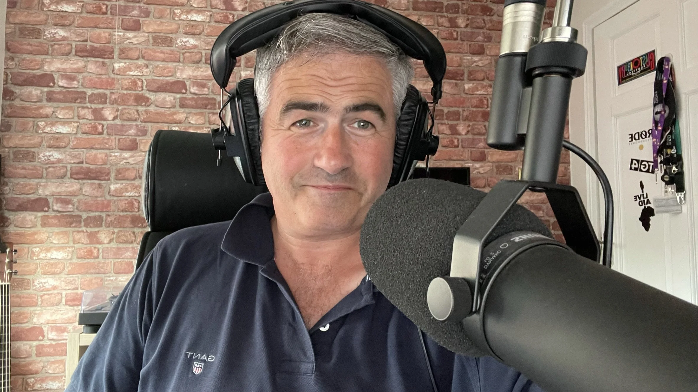 A man with gray hair and green eyes wearing a dark polo shirt with an American football team logo, sitting in front of a brick wall, and speaking into a large professional microphone, wearing large black headphones.