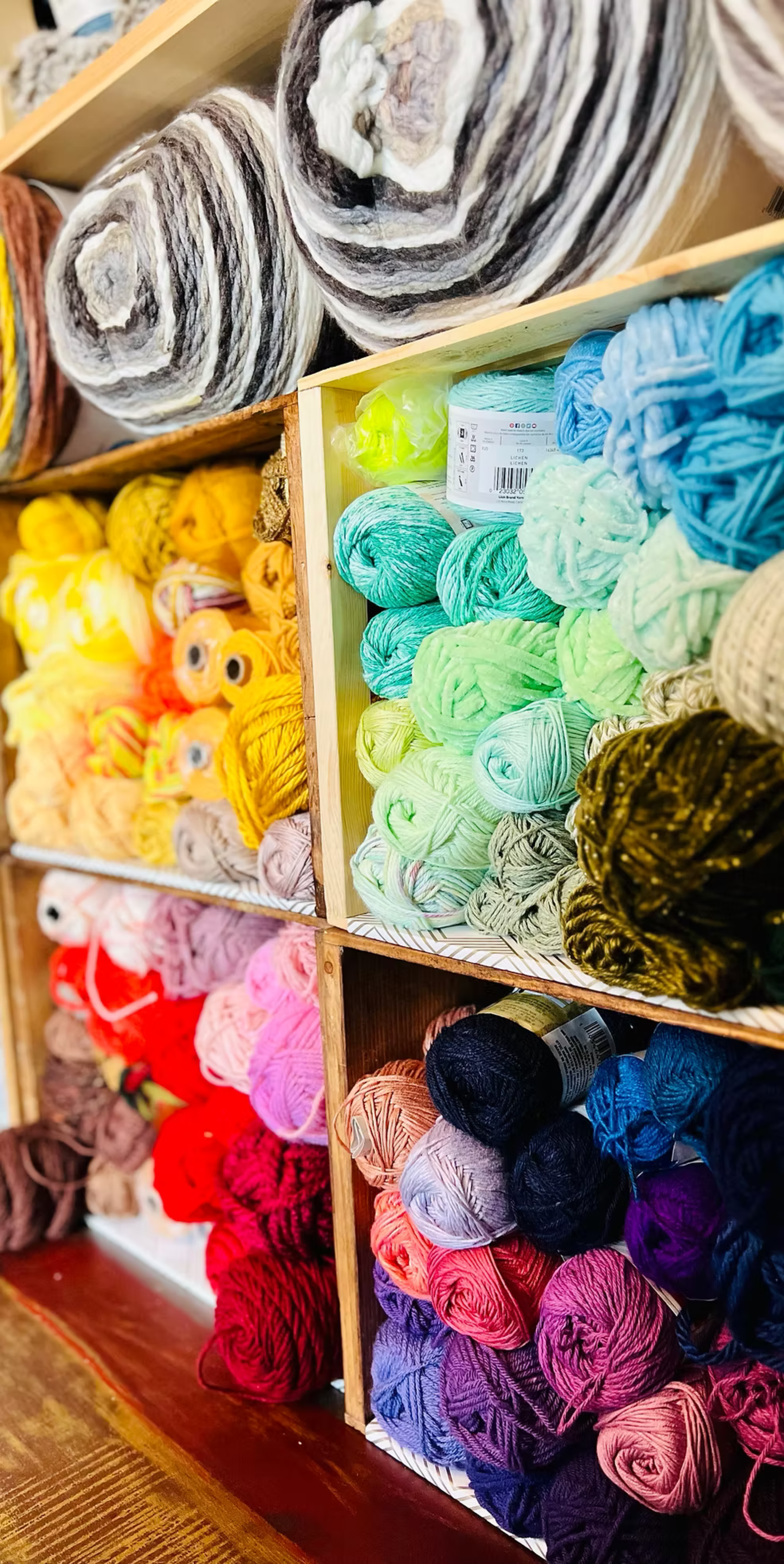 Colorful yarn skeins arranged on wooden shelves, with shades of black, white, gray, yellow, orange, pink, red, purple, and blue.