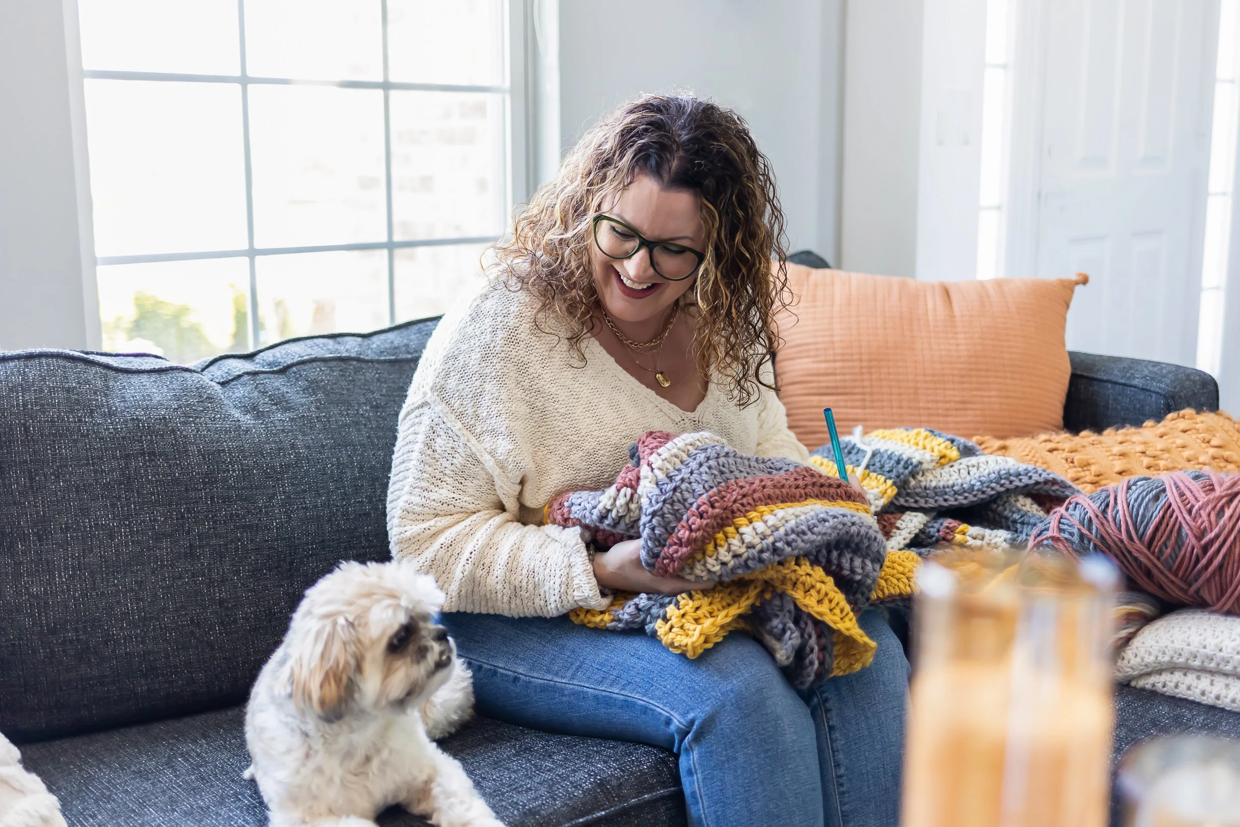 A woman with curly hair, glasses, and layered necklaces sitting on a dark gray couch, knitting colorful yarn, with a small white and brown puppy sitting on her lap. The room has large windows with white frames and a white door in the background.
