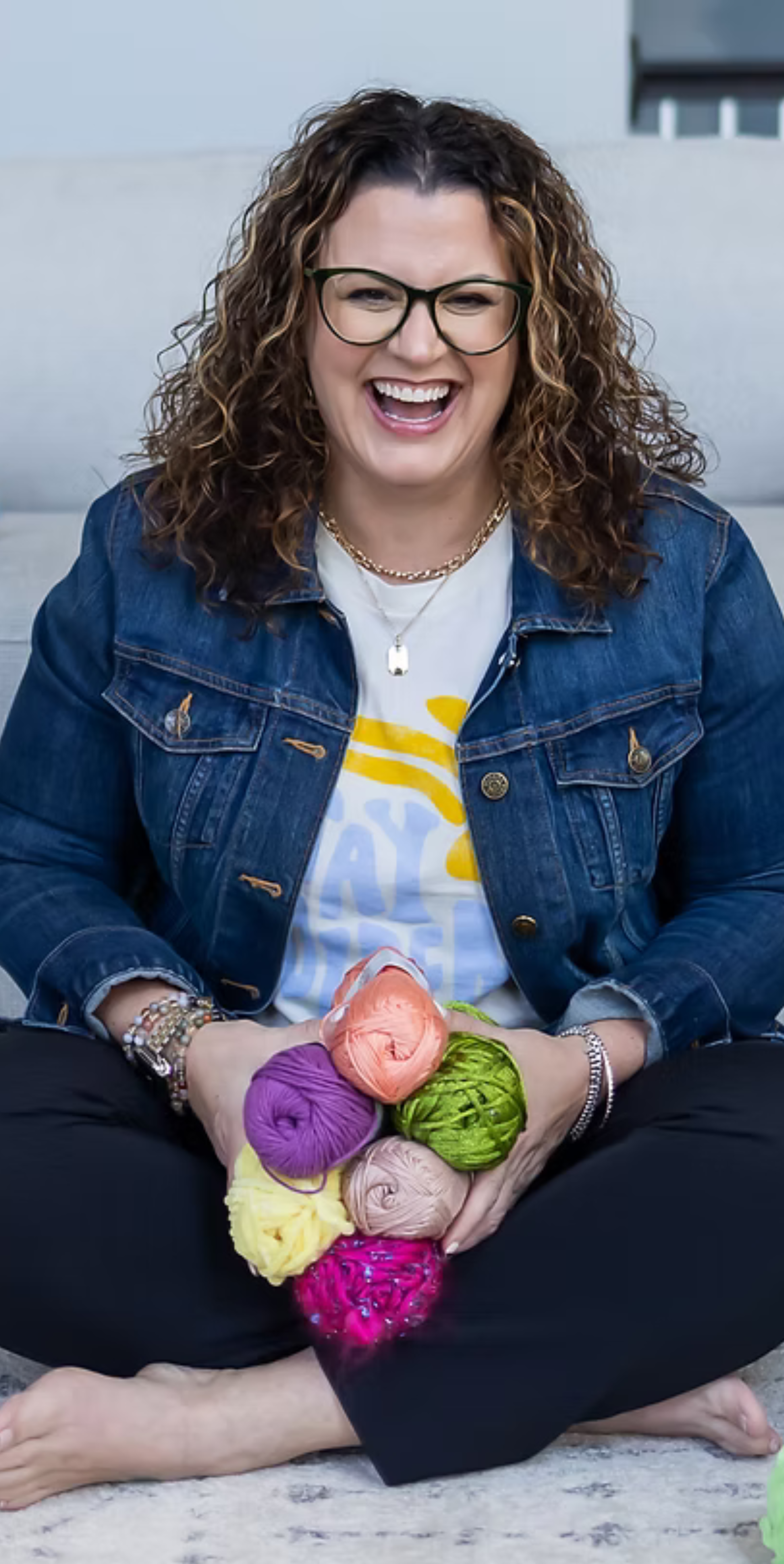 Woman with curly hair, glasses, and jewelry sitting on the floor with her legs crossed, holding multiple balls of colorful yarn.
