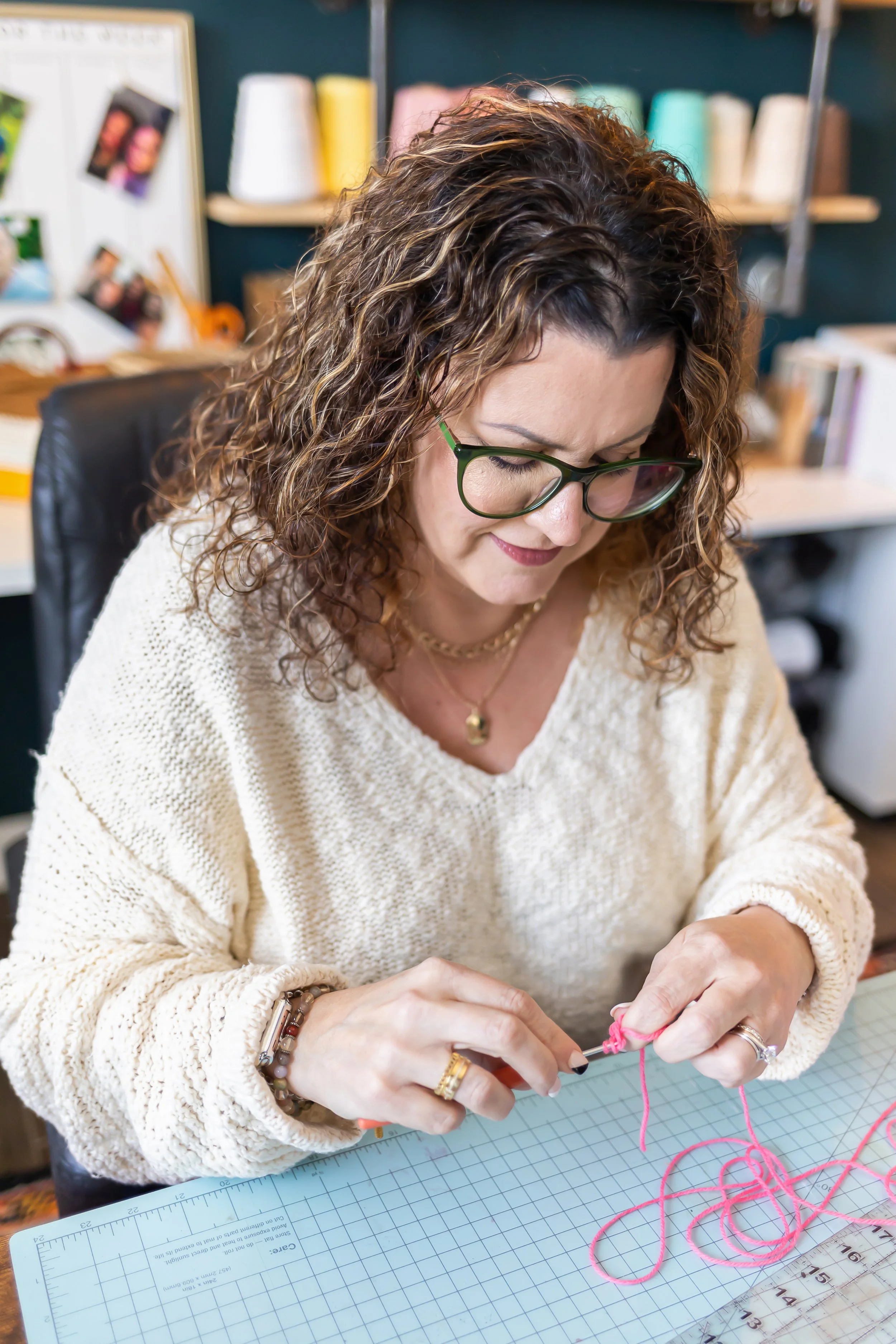 Woman with glasses and curly hair working on a craft project with pink yarn at a cutting mat in a craft room.