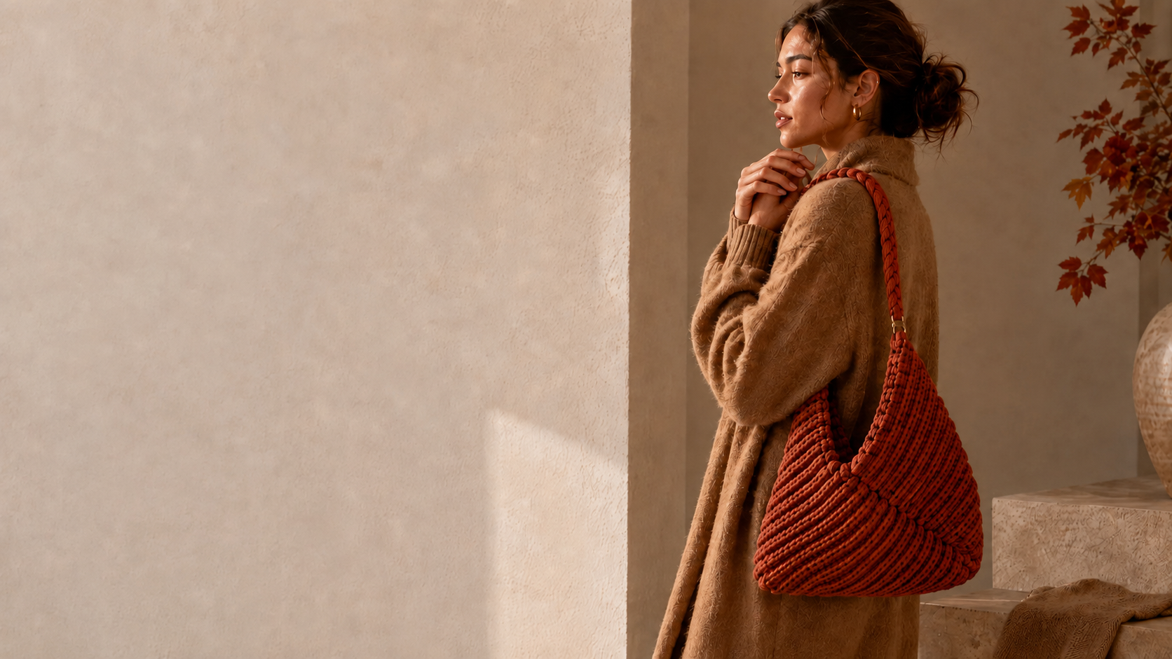 A woman with curly hair wearing a brown coat and holding a large, braided, orange handbag, standing next to a textured beige wall, with a vase and autumn leaves in the background.