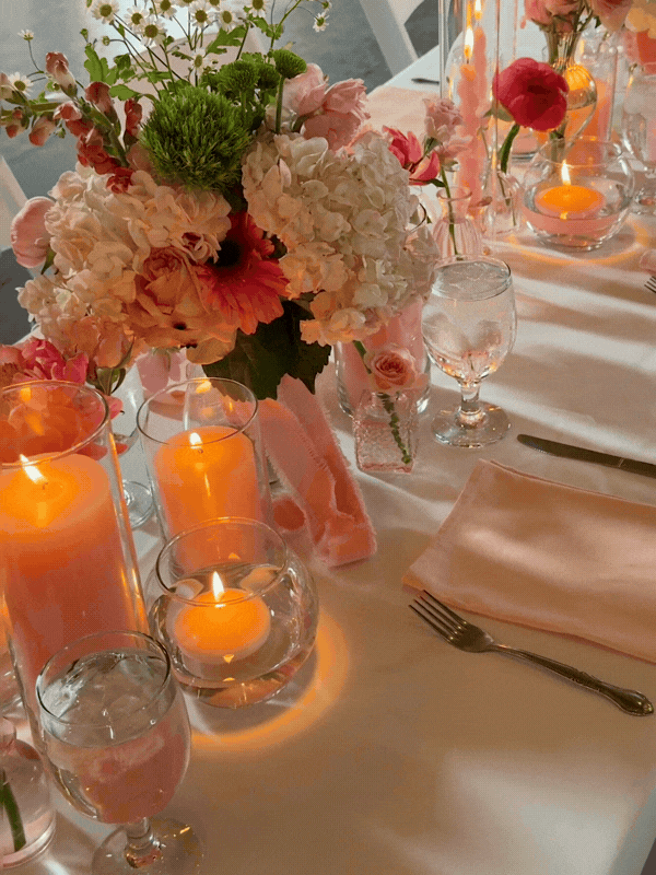A close-up of a table decorated with a bouquet of pink and white flowers, several lit candles in glass holders, and glassware including water glasses, set for a formal dinner.