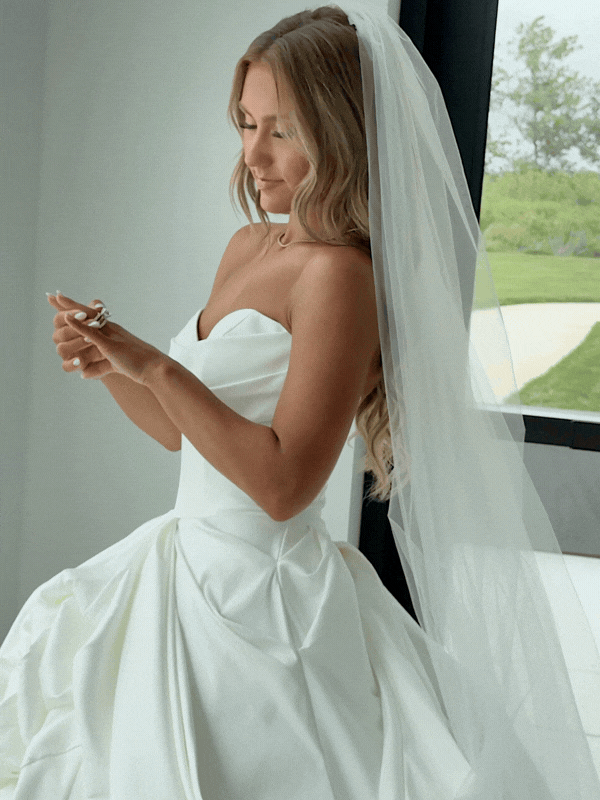 A woman in a white wedding dress with a veil is standing indoors, looking at her phone near a window with a green outdoor view.