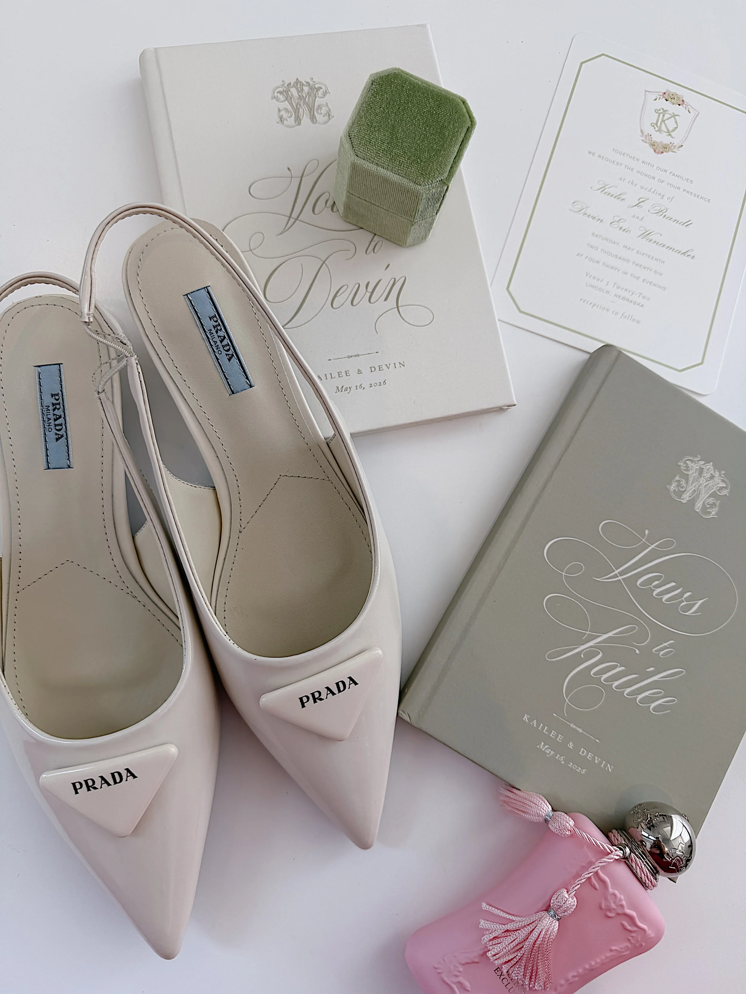 White Prada shoes, a green ring box, a white wedding invitation, a pink gift box with a tassel, and a gray book with elegant writing.