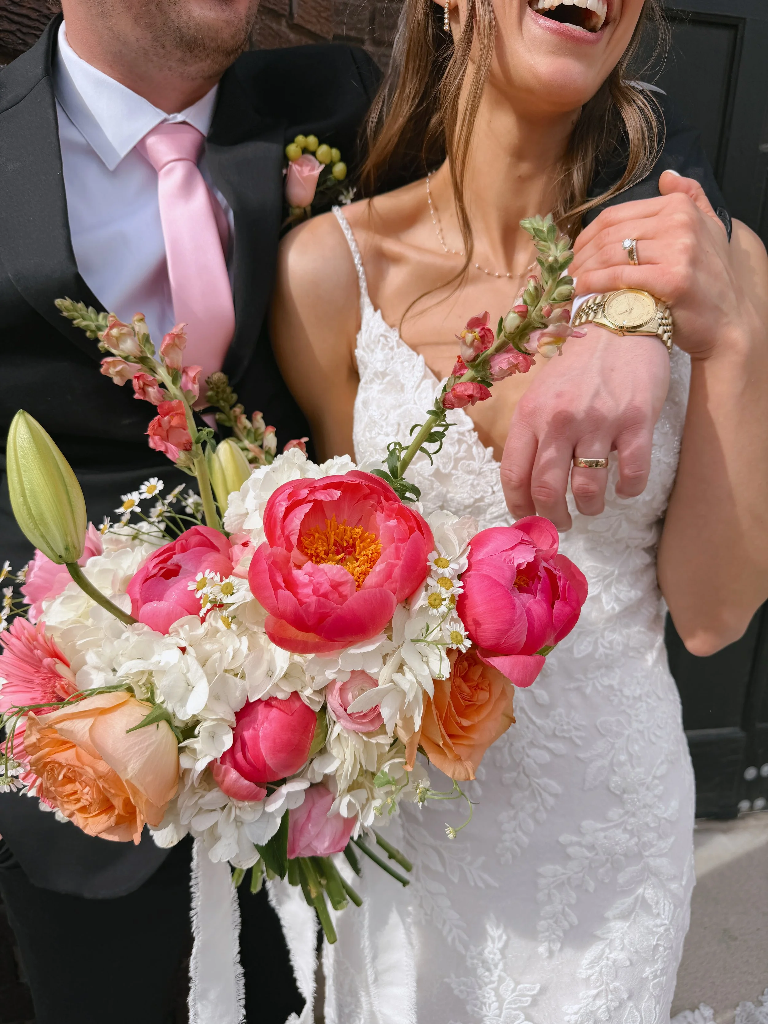 Close-up of a smiling bride and groom during their wedding, showing the bride's face, bouquet, and jewelry, with the groom in a suit with a pink tie and boutonniere.