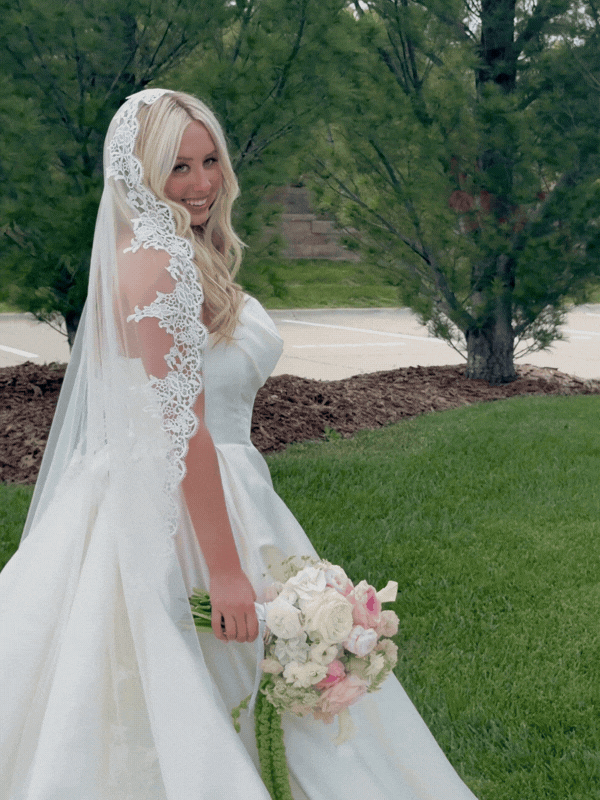 A smiling woman in a white wedding dress with lace details, holding a bouquet of pink and white flowers, standing outdoors on green grass with trees in the background.