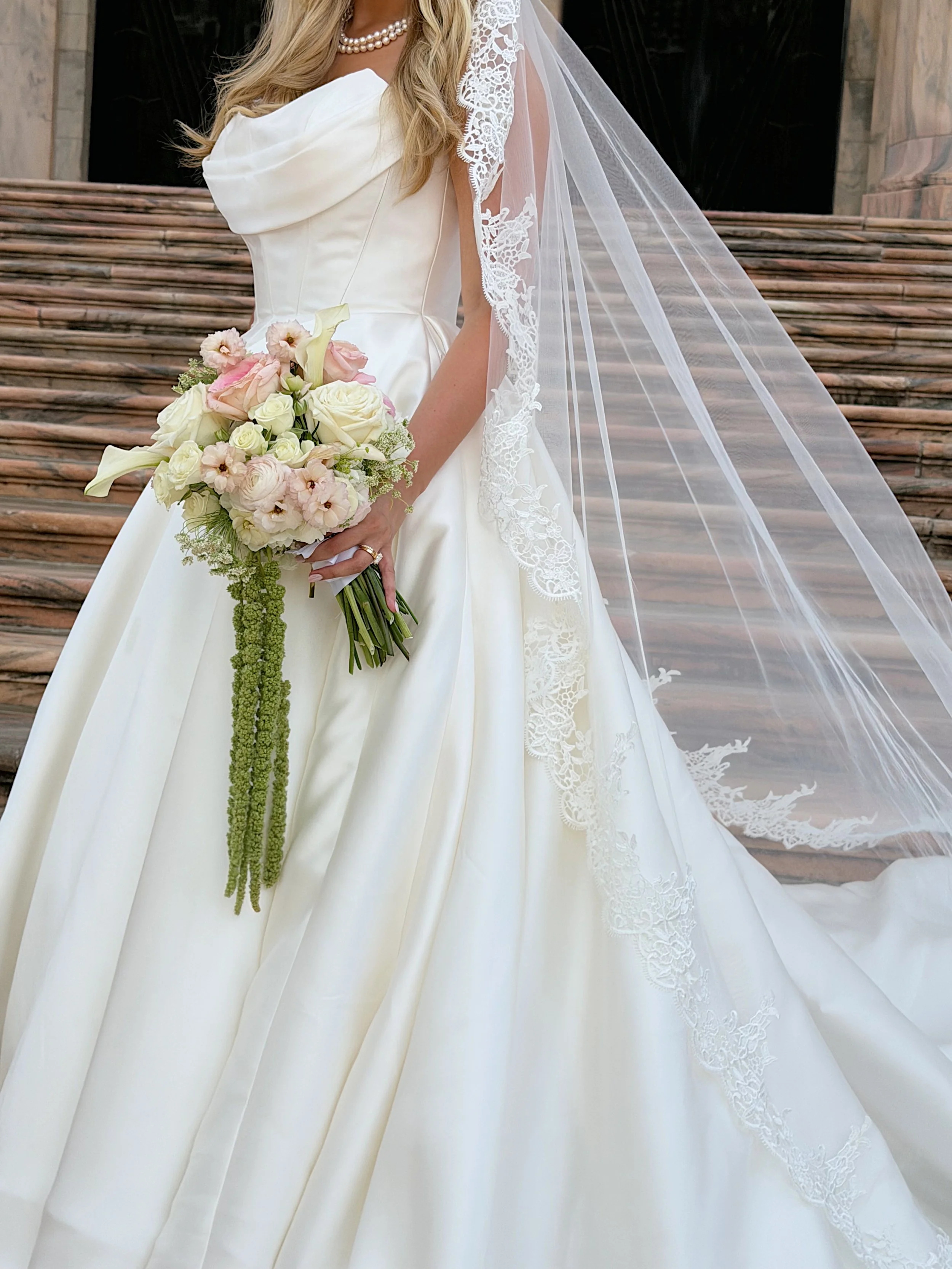 Bride wearing a white wedding gown holding a bouquet of white and pink flowers, with lace details on her veil, standing in front of stone steps.