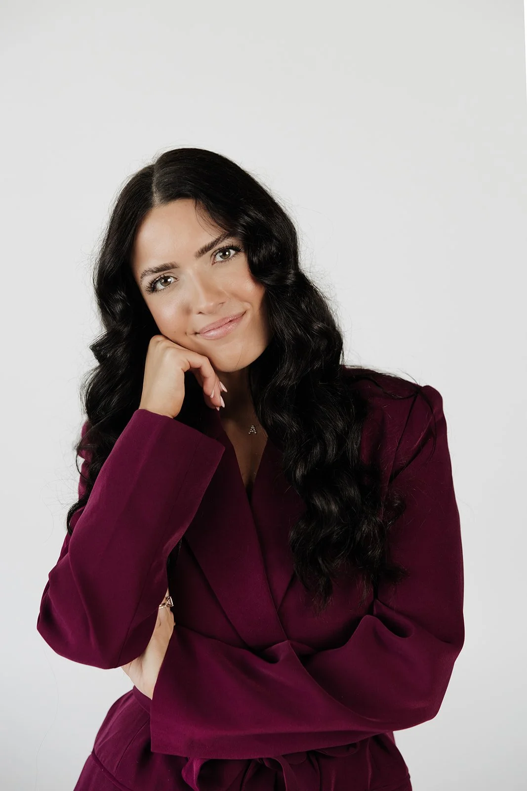 Woman with long black wavy hair, wearing a burgundy blazer, posing with her chin resting on her hand, smiling softly against a white background.