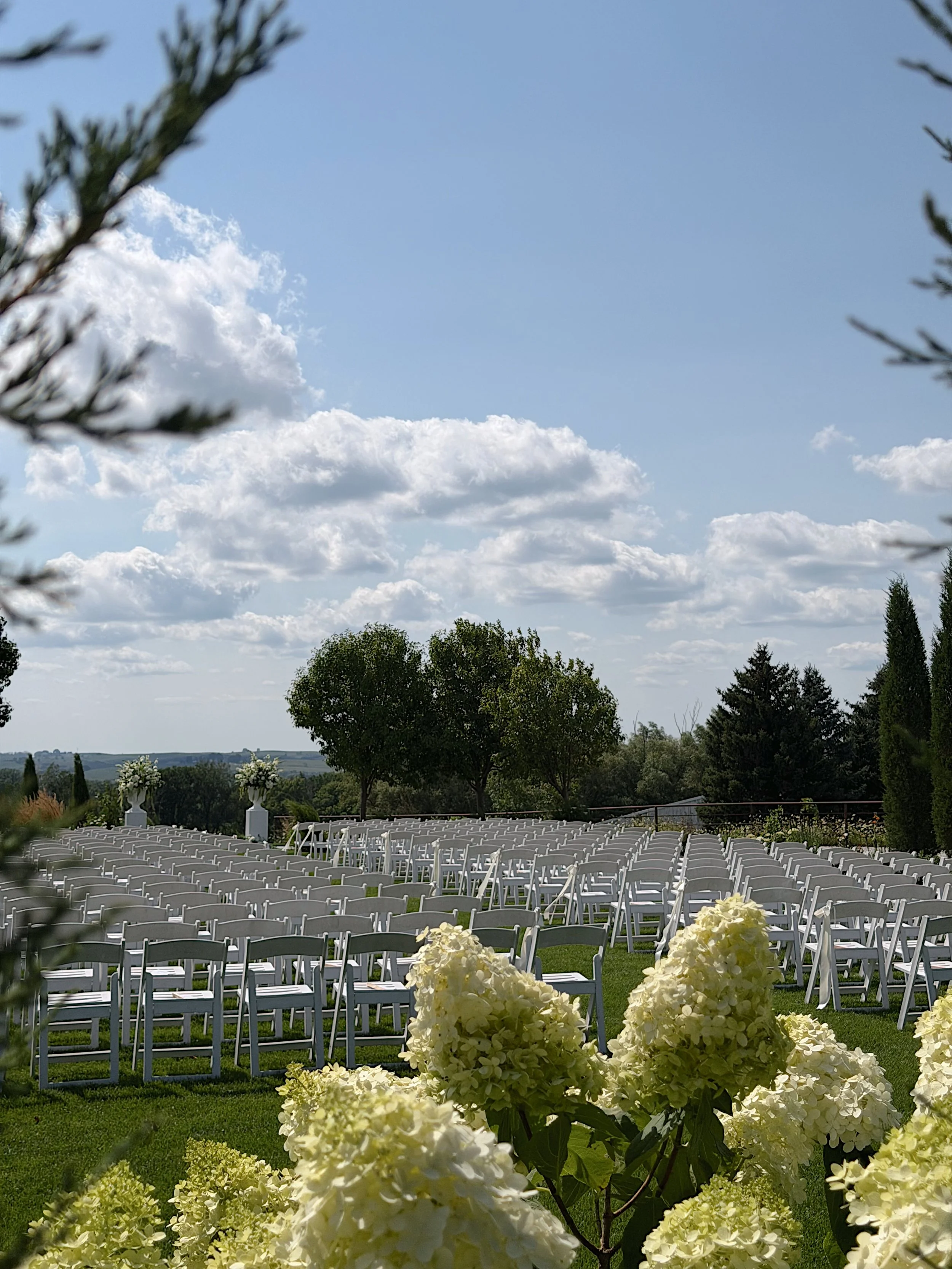 Outdoor wedding ceremony setup with numerous white chairs arranged in rows, decorated with large white flower arrangements, lush green trees, and partly cloudy sky.