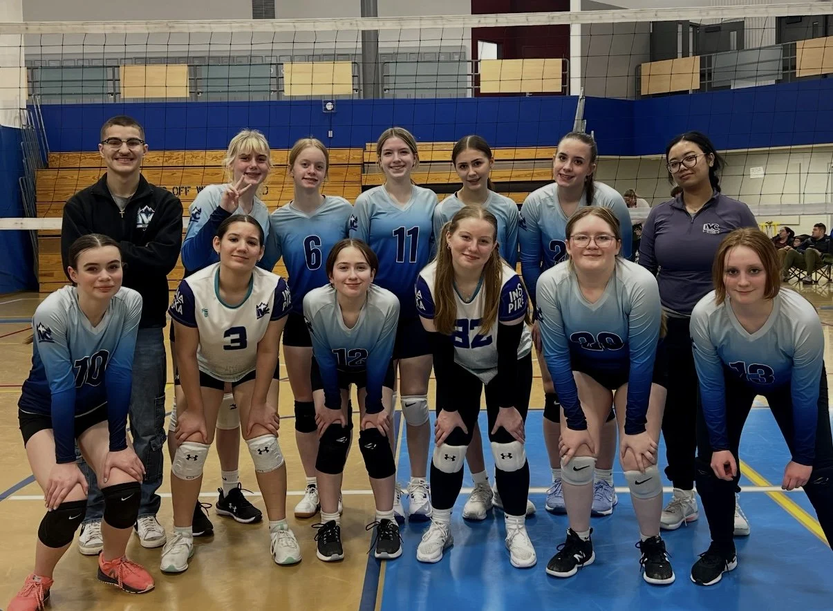 A girls' volleyball team posing for a group photo in a gymnasium, with a net behind them and a blue and yellow colored background, along with their coaches.