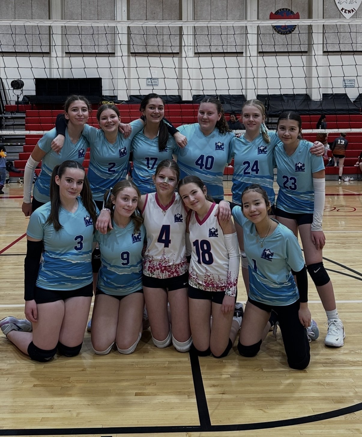 A volleyball team of 11 young women in blue and white uniforms posing indoors on a gymnasium floor with a volleyball net behind them.