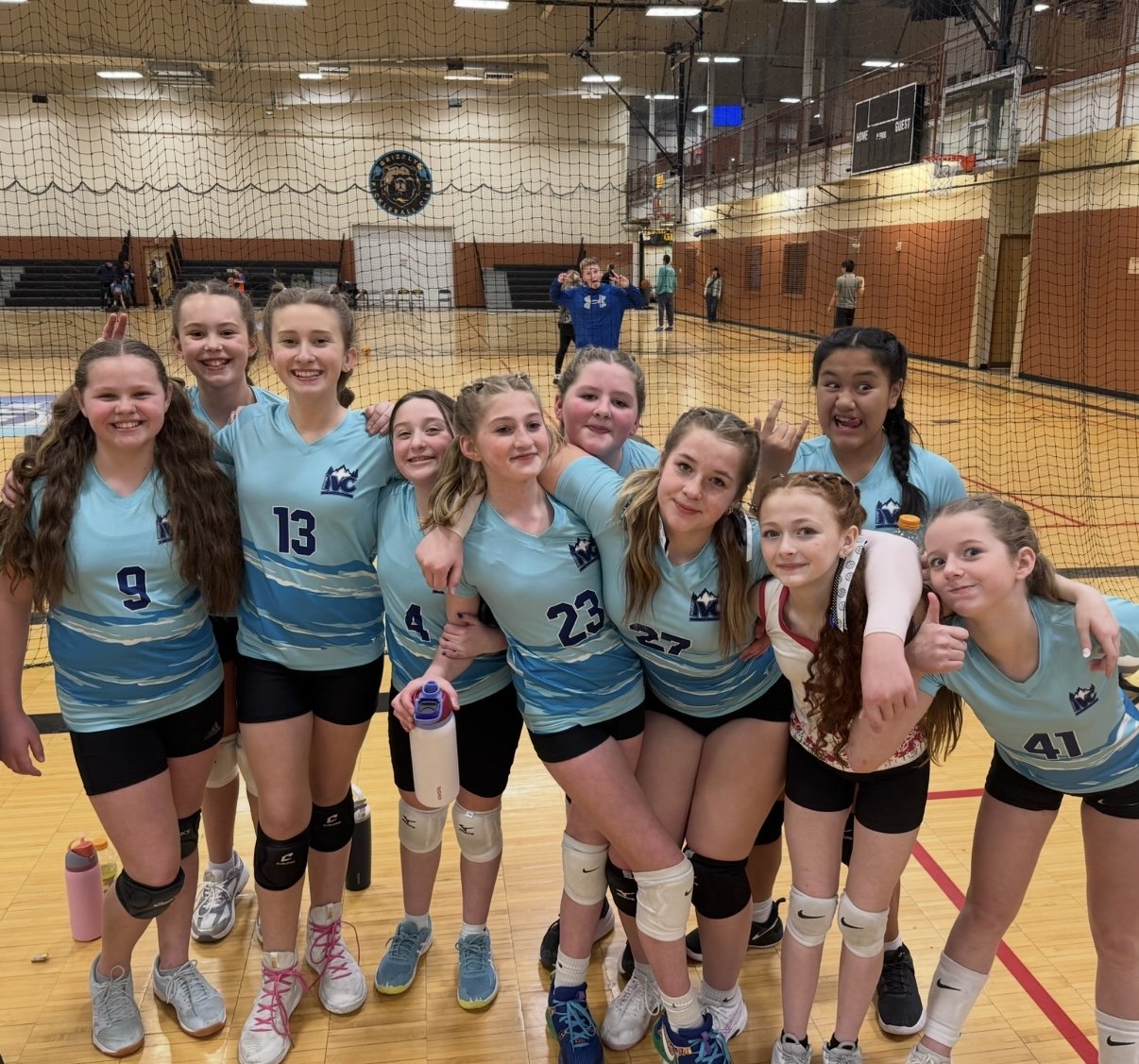 A group of young female volleyball players in blue uniforms with numbers 9, 13, 4, 23, 27, 41, and others, posing together on an indoor court. They are smiling and show camaraderie, with some fans and players in the background.