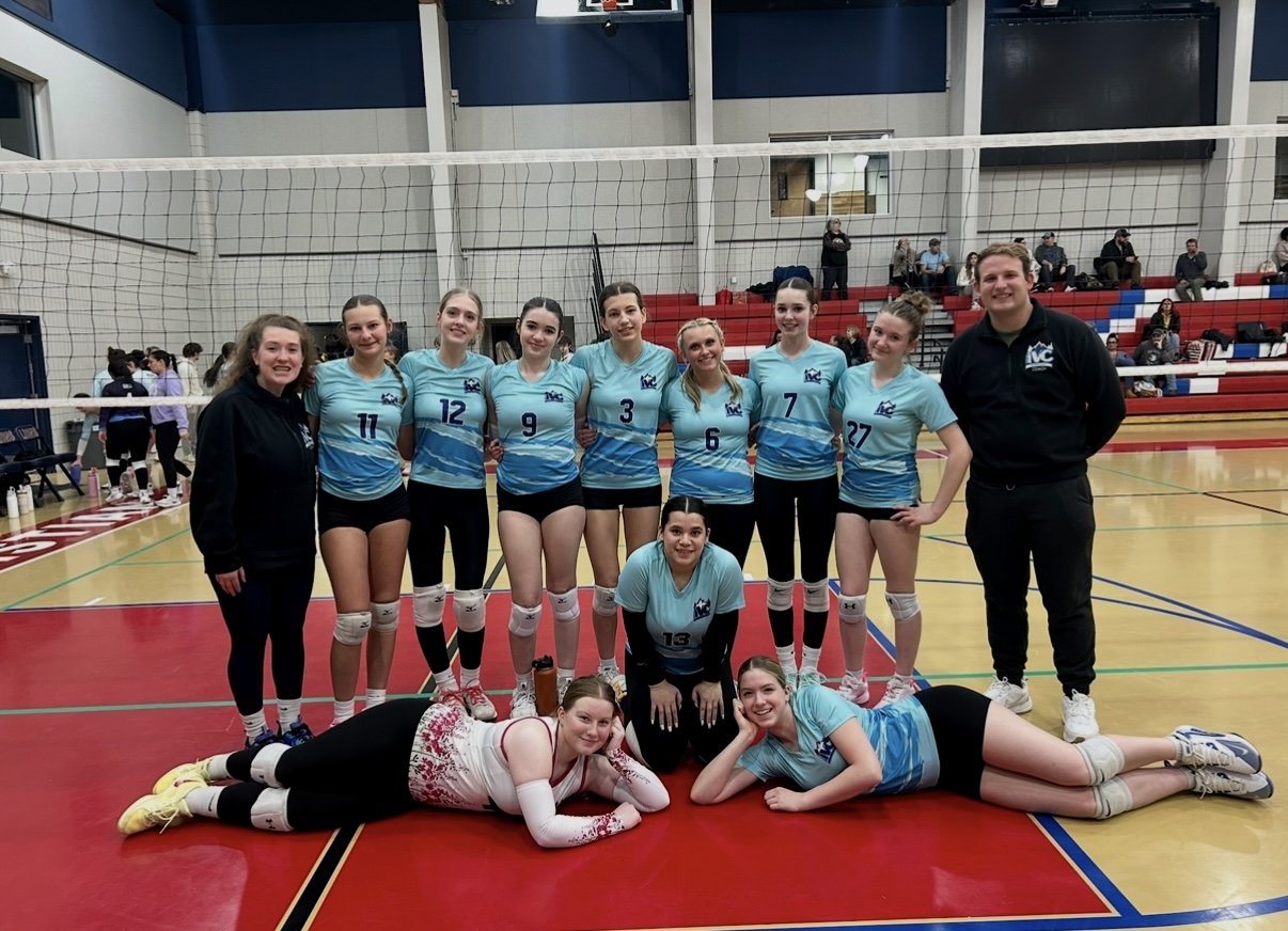 A volleyball team consisting of nine girls and two coaches posing in a gymnasium. The team is wearing matching blue jerseys with black shorts, with players' numbers visible. Two players are lying on the floor in front, and the rest standing behind. The coaches are at either end of the group, one in a black jacket and the other in black pants and a black top. There are spectators seated on red bleachers in the background.