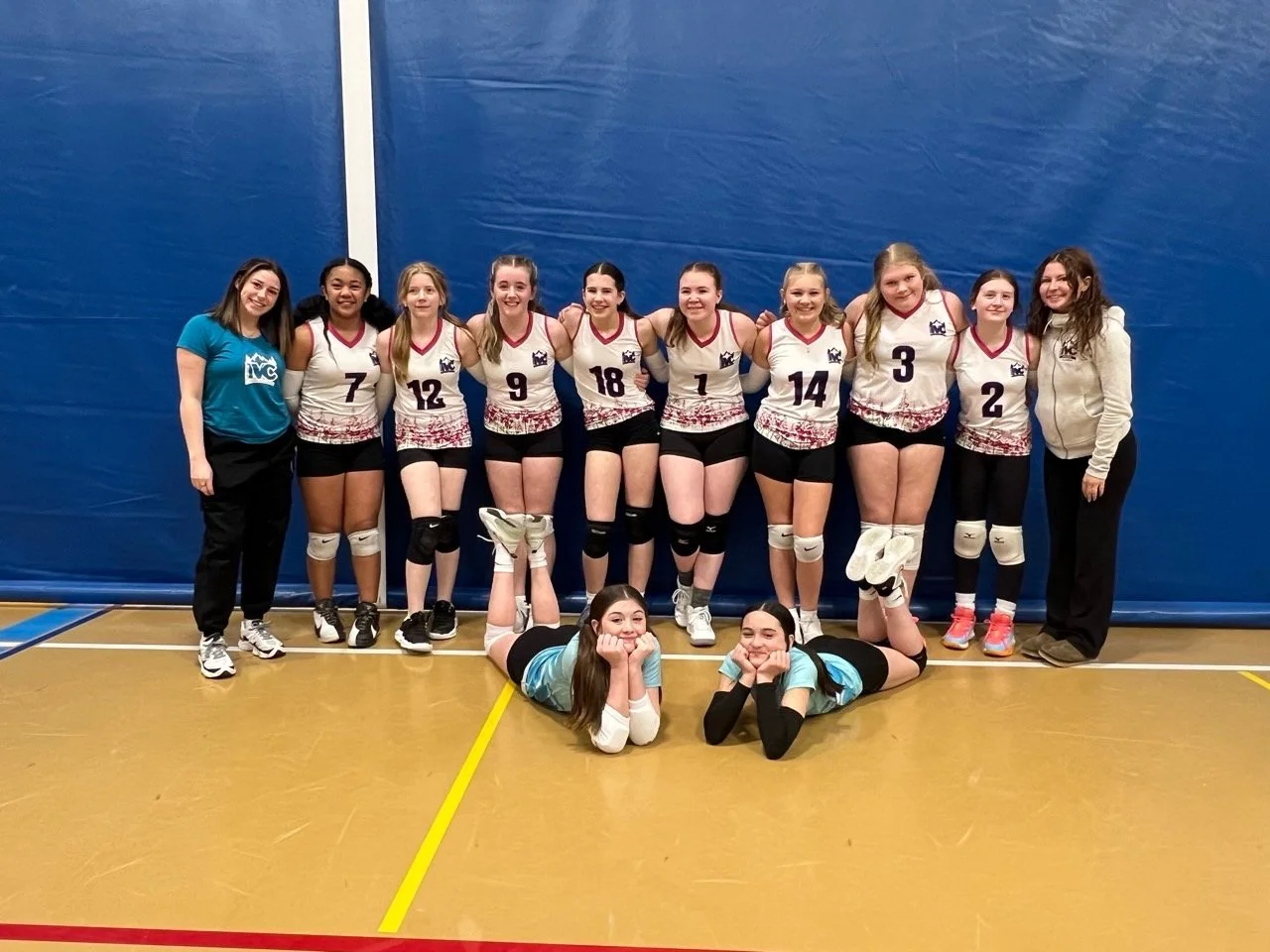 A girls' volleyball team with ten players in uniform and two coaches posing together in a gymnasium, with two girls lying in front.