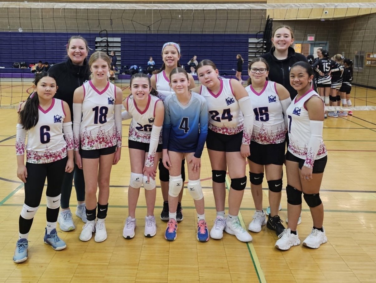 A youth volleyball team with two coaches posing together on a gymnasium court. The team members are wearing matching uniforms with numbers, and there are volleyball nets and other players in the background.