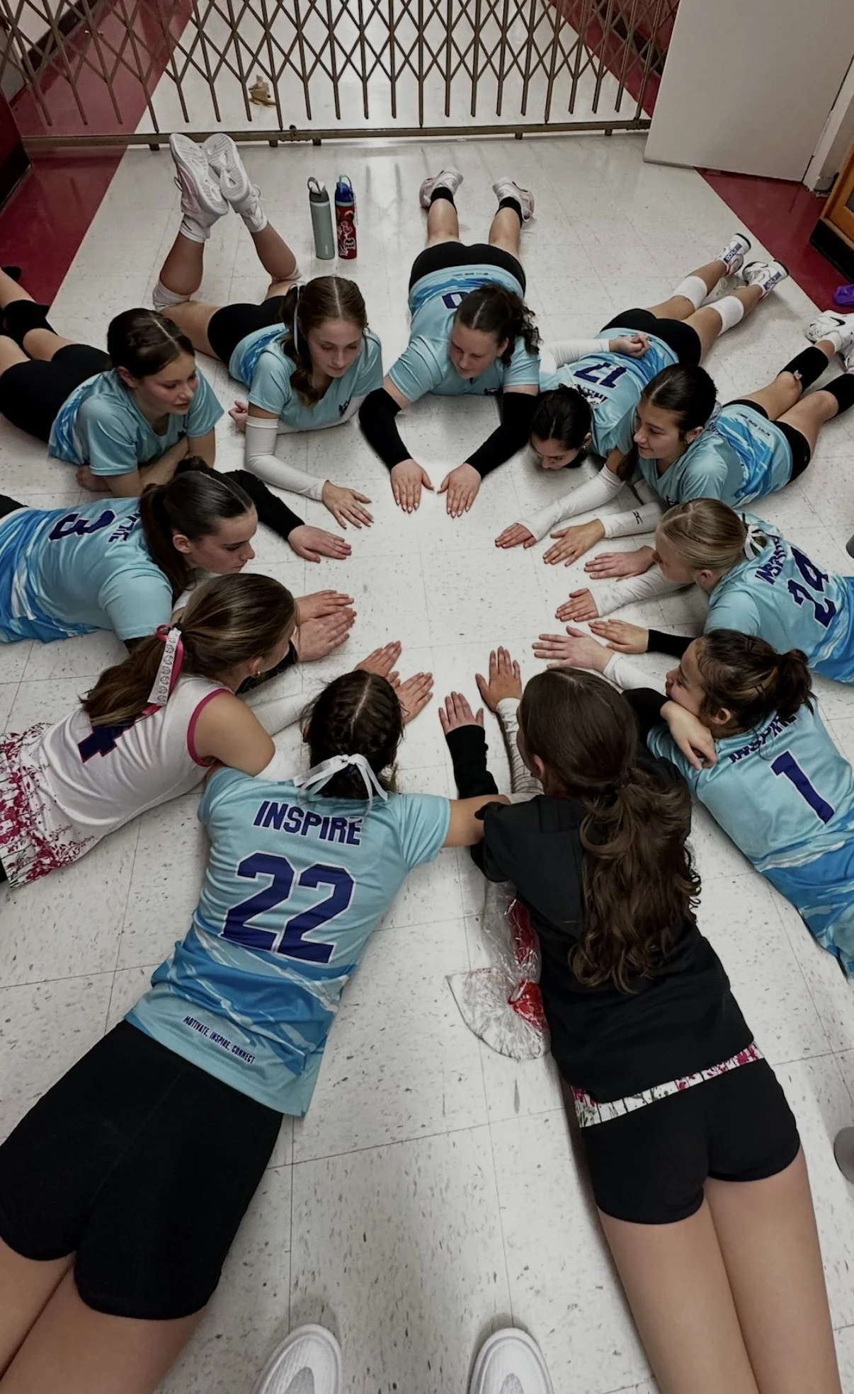 A group of young girls in sports uniforms forming a circle on the floor, lying on their stomachs with their hands extended towards the center, engaging in a team-building activity or cheer.