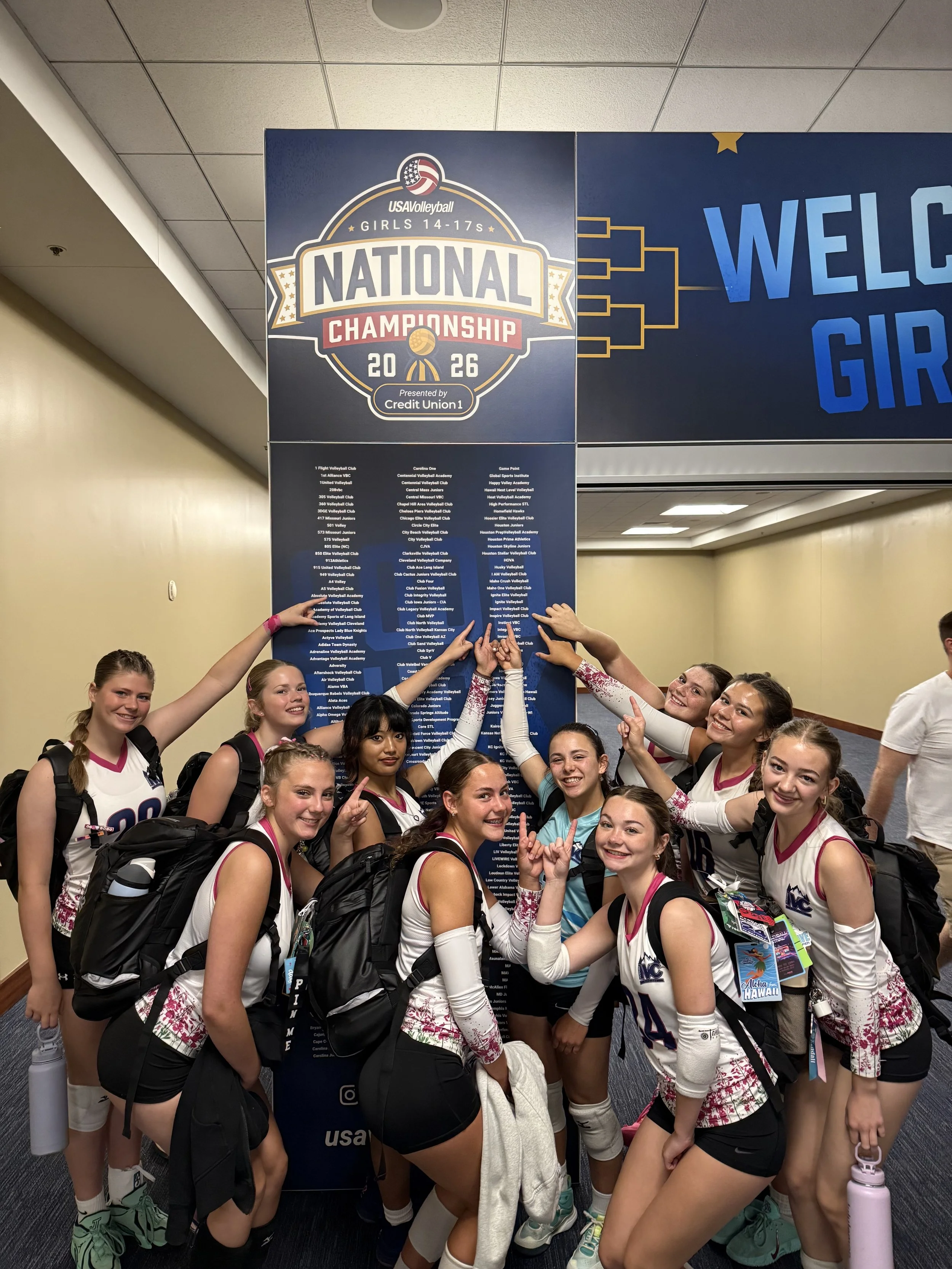 A group of young female volleyball players in athletic uniforms standing together at an indoor sports venue, smiling and gesturing at a large sign for the 2026 USA Volleyball Girls' National Championship.