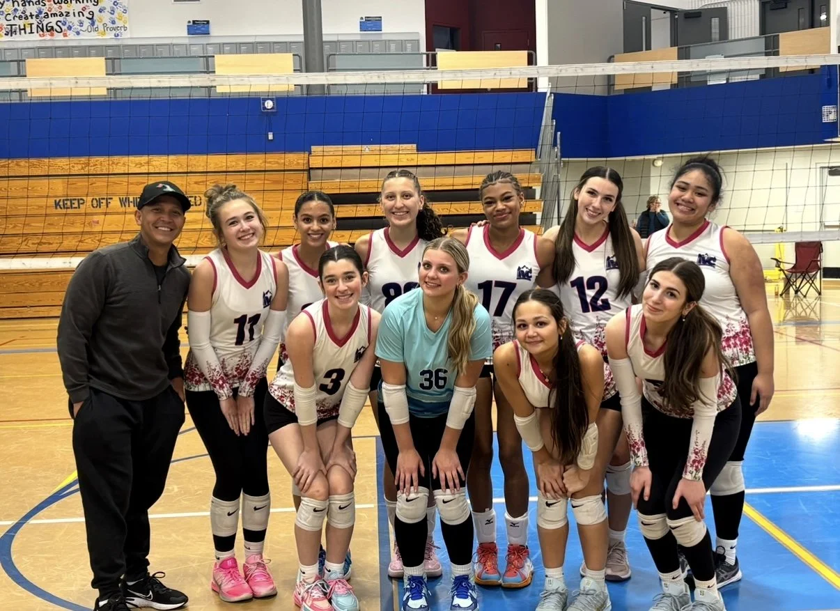 A girls' volleyball team with their coach in a gymnasium, posing for a group photo after a game. The team is wearing white and pink uniforms, and the gym has wooden and blue bleachers in the background.