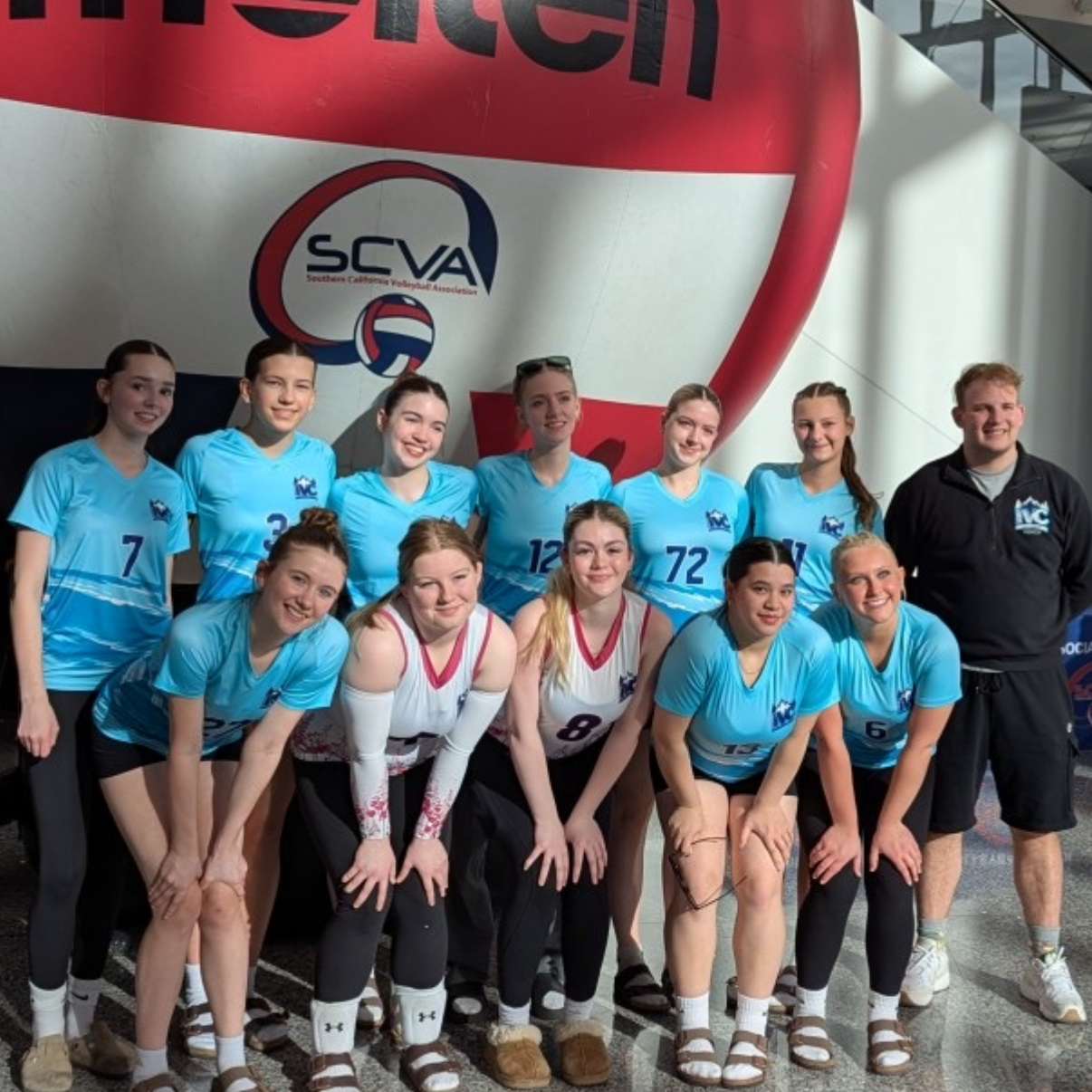 Group of young female volleyball players in light blue jerseys and a coach in a black jacket, standing indoors in front of a large banner with the Southern California Volleyball Association (SCVA) logo.