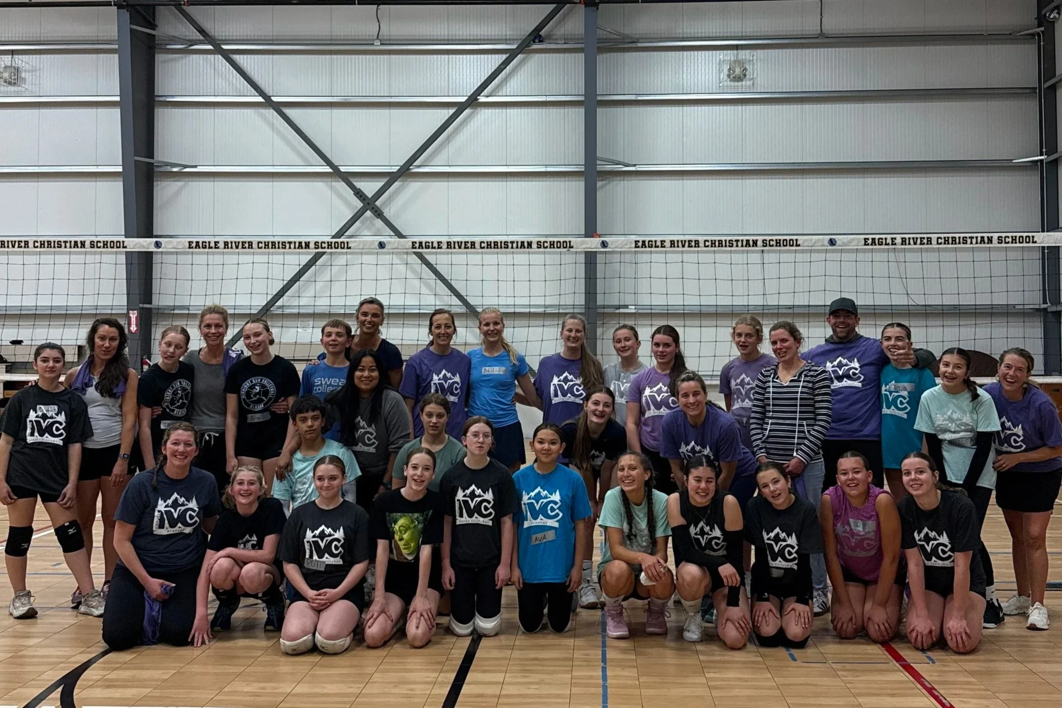 Group of young women and girls in athletic uniforms posing in a gymnasium with a volleyball net behind them.