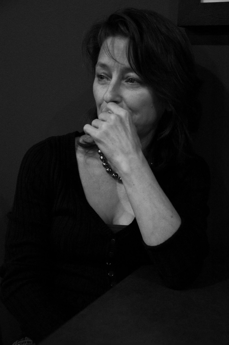 A black-and-white photo of Tamara Skidmore, writer, with medium-length dark hair, wearing a dark top with a button-up front and a necklace, sitting at a table with her hand covering her mouth, looking thoughtful or pensive.