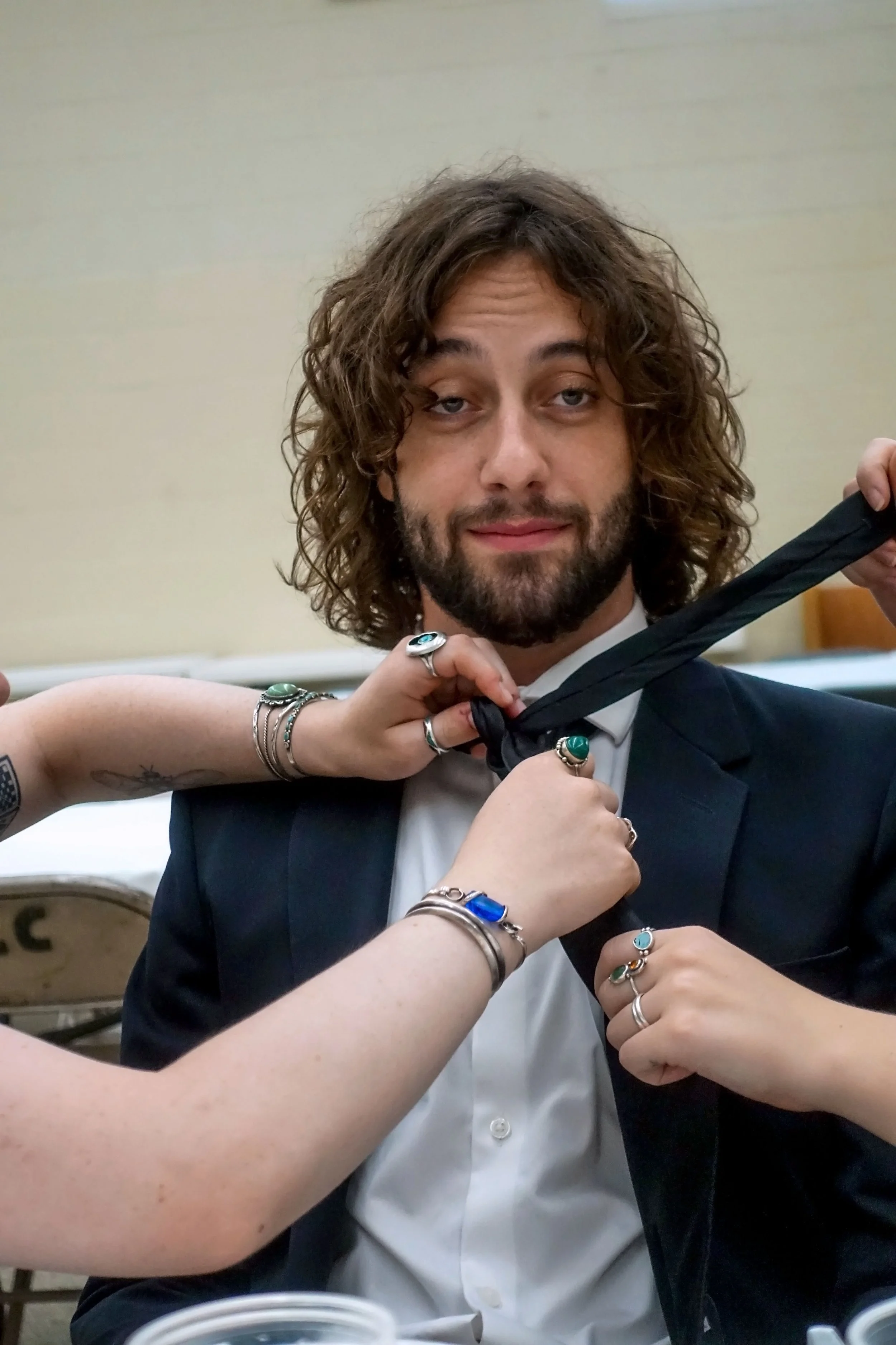 A man, Tamara Skidmore's son Eliot, with long curly hair and a beard, dressed in a tuxedo, is holding a black bow tie. Multiple hands with jewelry are adjusting the tie around his neck.